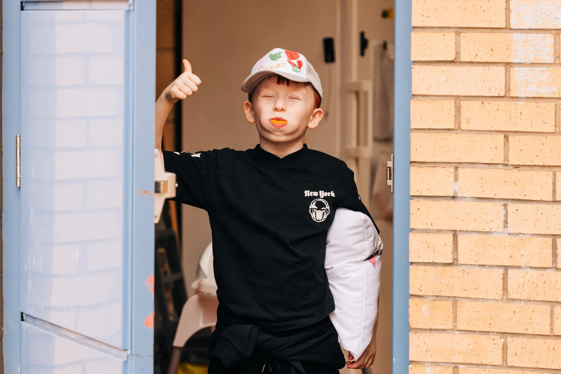 A young boy is standing at a doorway, giving a thumbs-up, with his eyes closed, and a thoughtful expression on his face. He is holding a white pillow in his left arm and wearing a baseball cap and a black t-shirt that has the words 'New York' and a graphic of headphones on it.