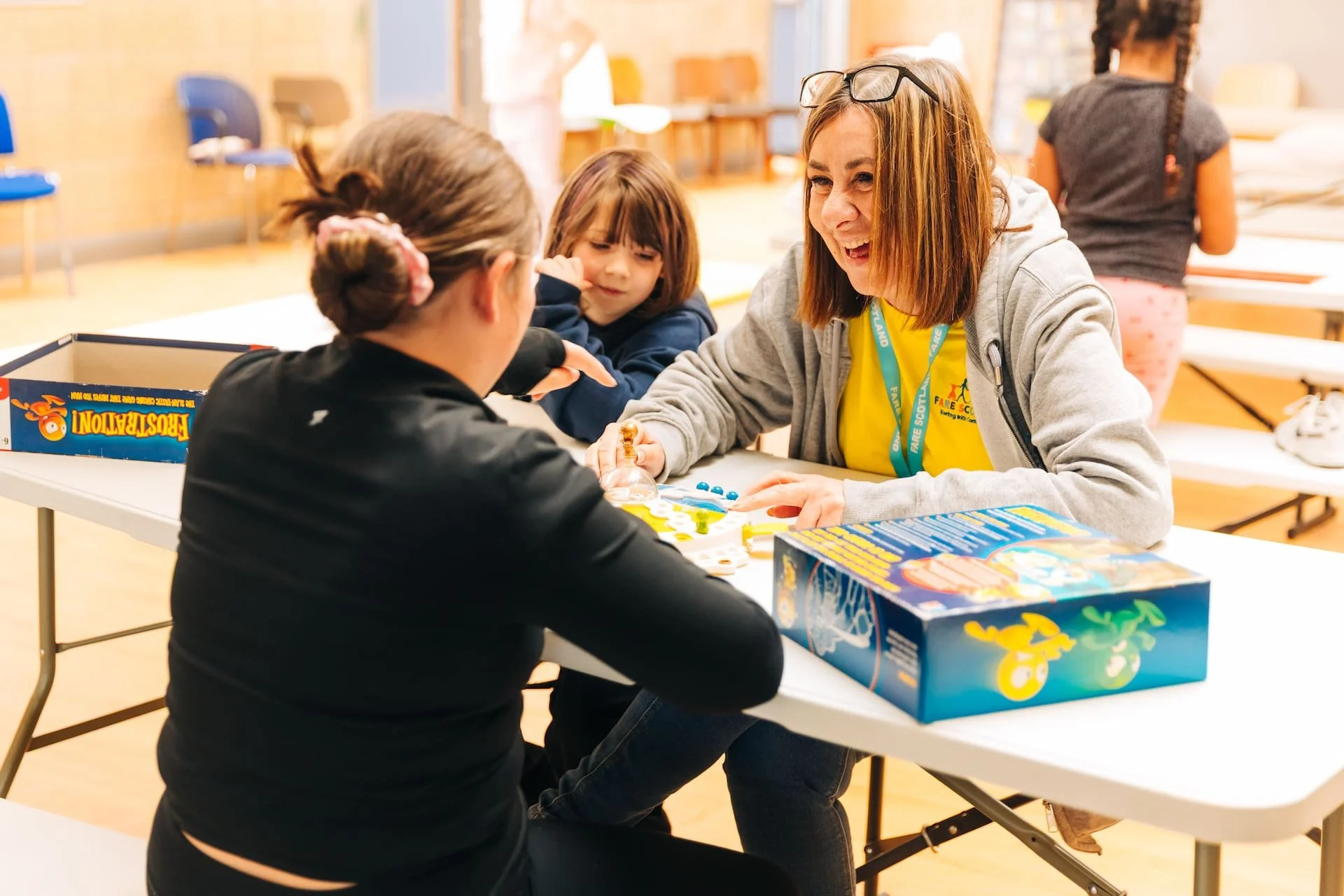 A woman playing a board game with two young girls in a classroom setting, with a third girl in the background, all seated at a white table.