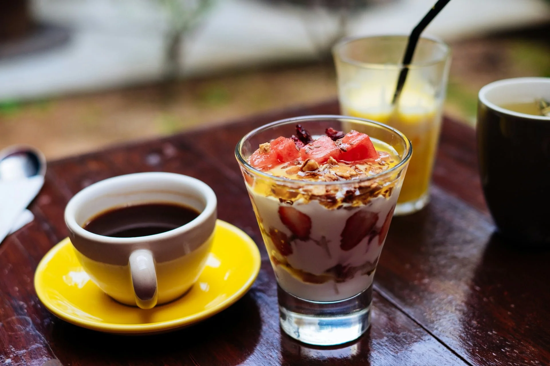 A glass of layered yogurt with strawberries, granola, and honey, a cup of black coffee on a yellow saucer, a glass of orange juice with a black straw, and a cup of tea on a wooden table.
