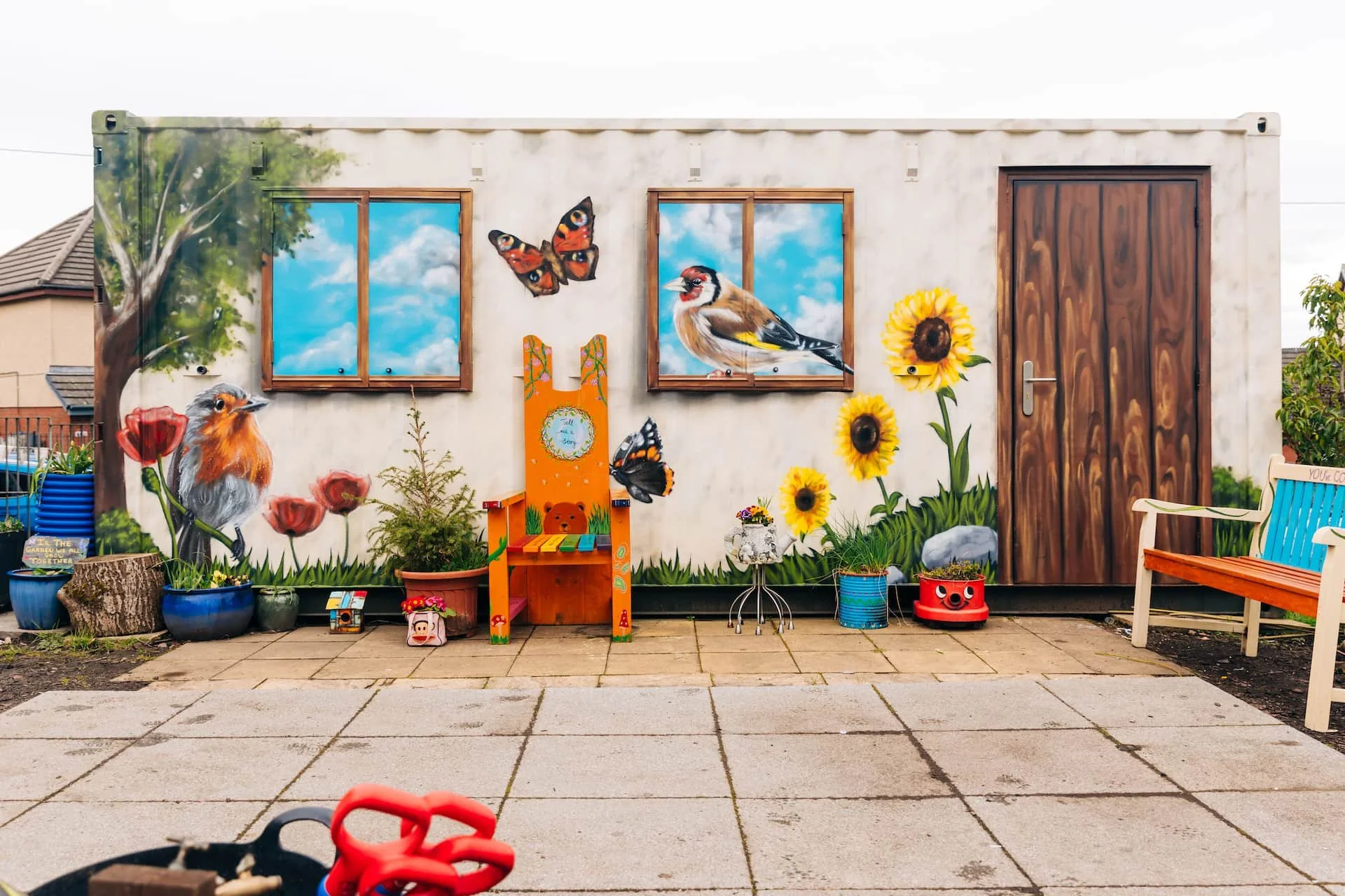 Colorful mural of birds, butterflies, and flowers on a white wall outside a house with a wooden door and a window showing a sky with clouds. Children's toys and plants are in front.