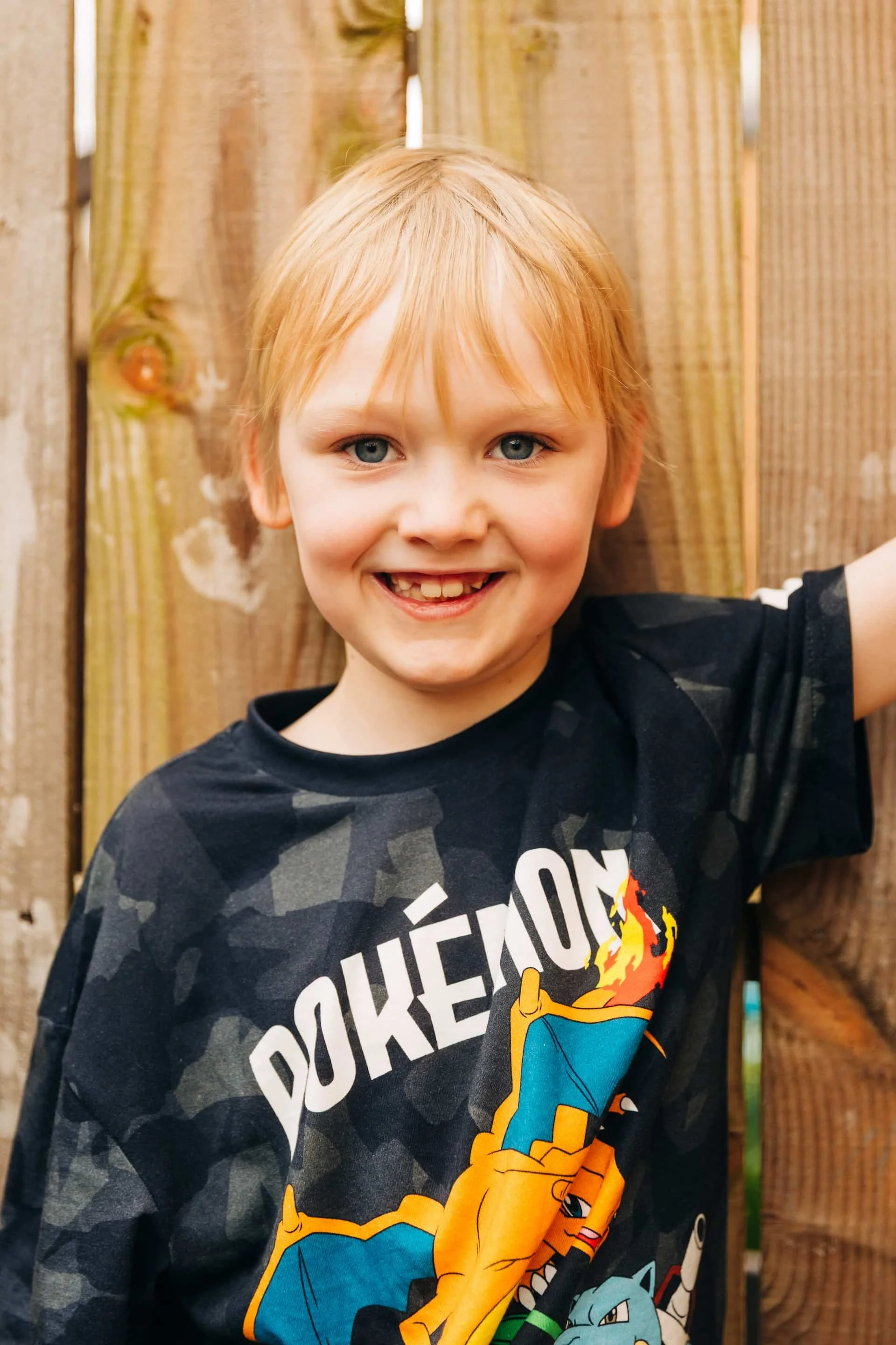 A young boy with blonde hair and blue eyes smiling outdoors, wearing a black Fortnite-themed t-shirt, standing in front of a wooden fence.
