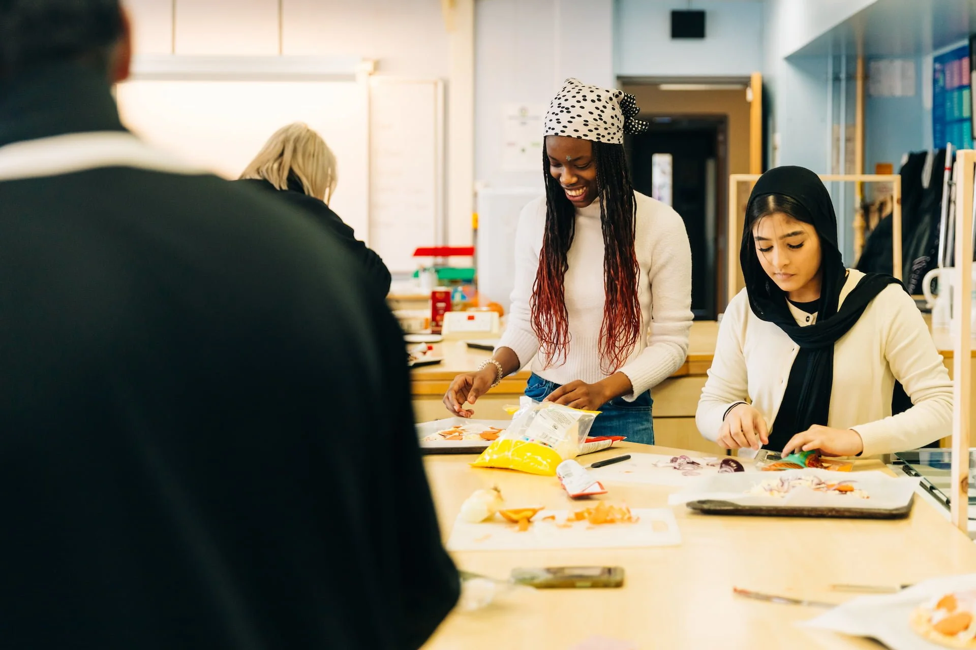 People in a cooking class preparing food at a countertop, some wearing headscarves, one woman smiling and working with ingredients.