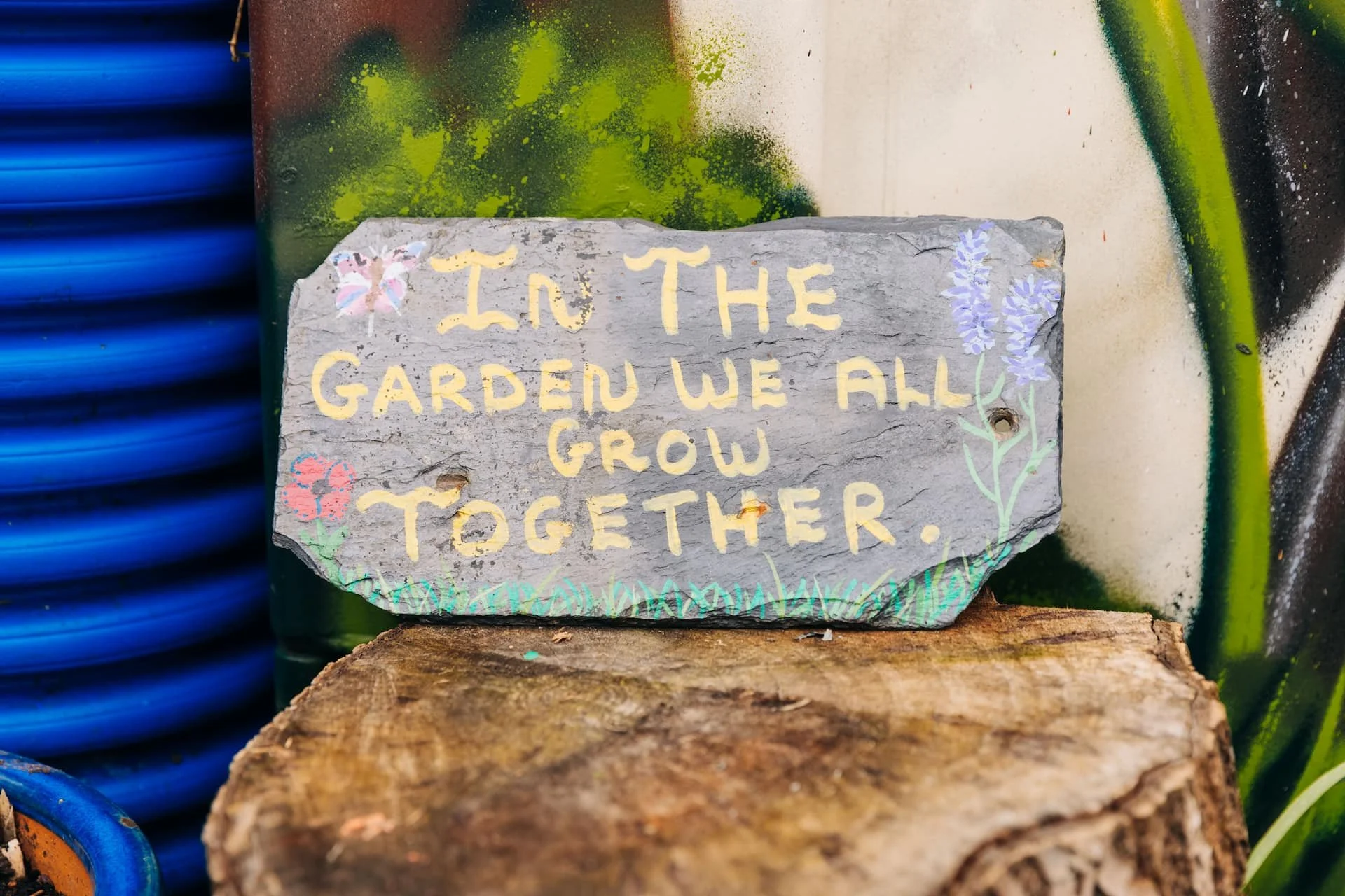 A painted stone with the phrase "In the garden we all grow together" surrounded by colorful flower illustrations, placed on a wooden surface.