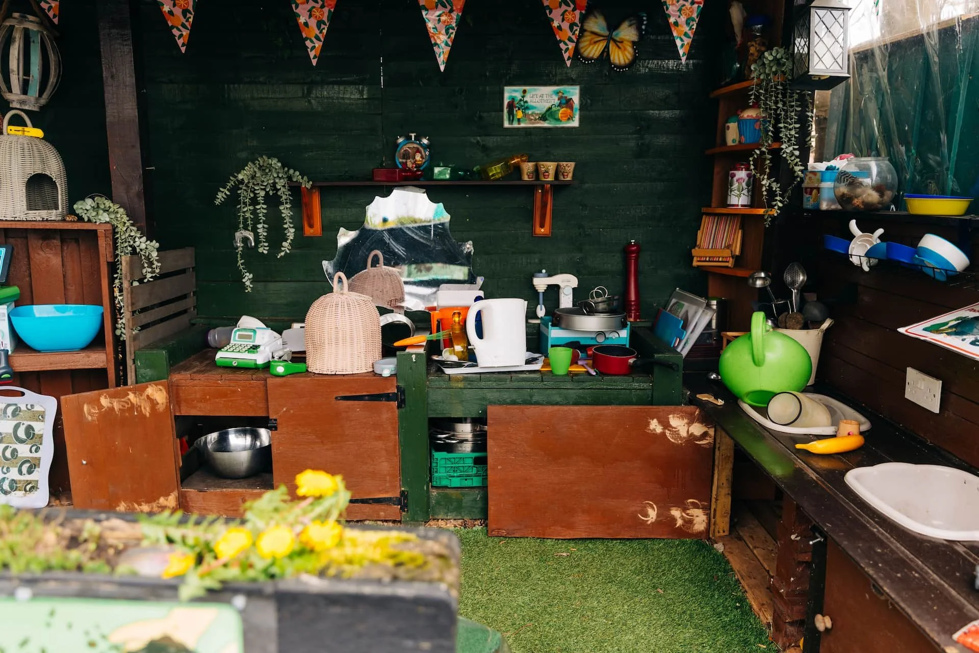 Messy outdoor kitchen with green and wooden walls, cluttered countertop with pots, pans, a knife, a green watering can, a white plastic bowl, a yellow pepper, and other kitchen items. Decorative butterfly and bunting hang above, plants and shelves wi