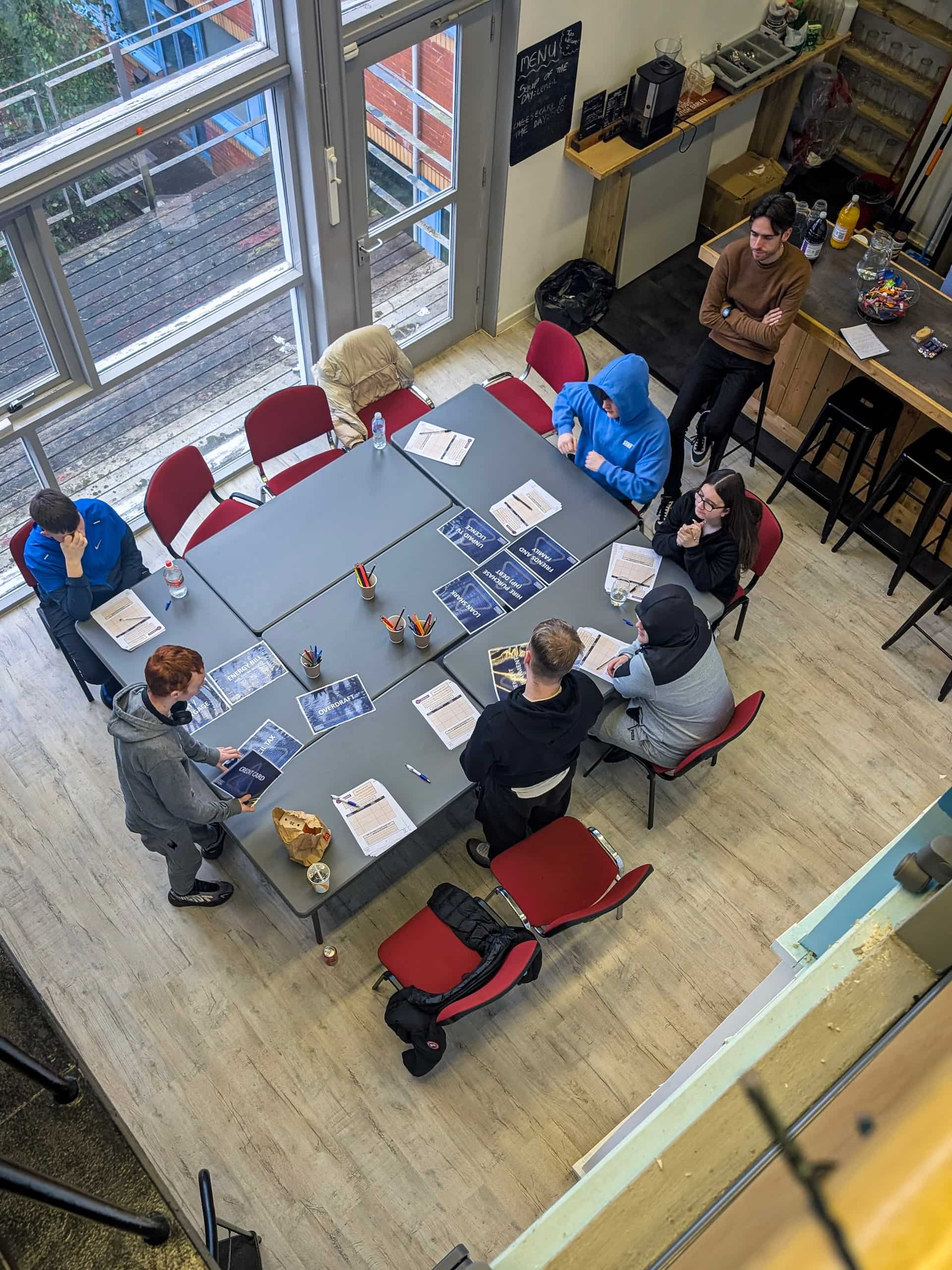 Group of eight people gathered around a rectangular table in a casual meeting room, with some reading materials, water bottles, and snacks. Four people are seated, two are standing, and another person stands in the background near a bar area with bottles and supplies. Large windows and a sliding glass door provide natural light.