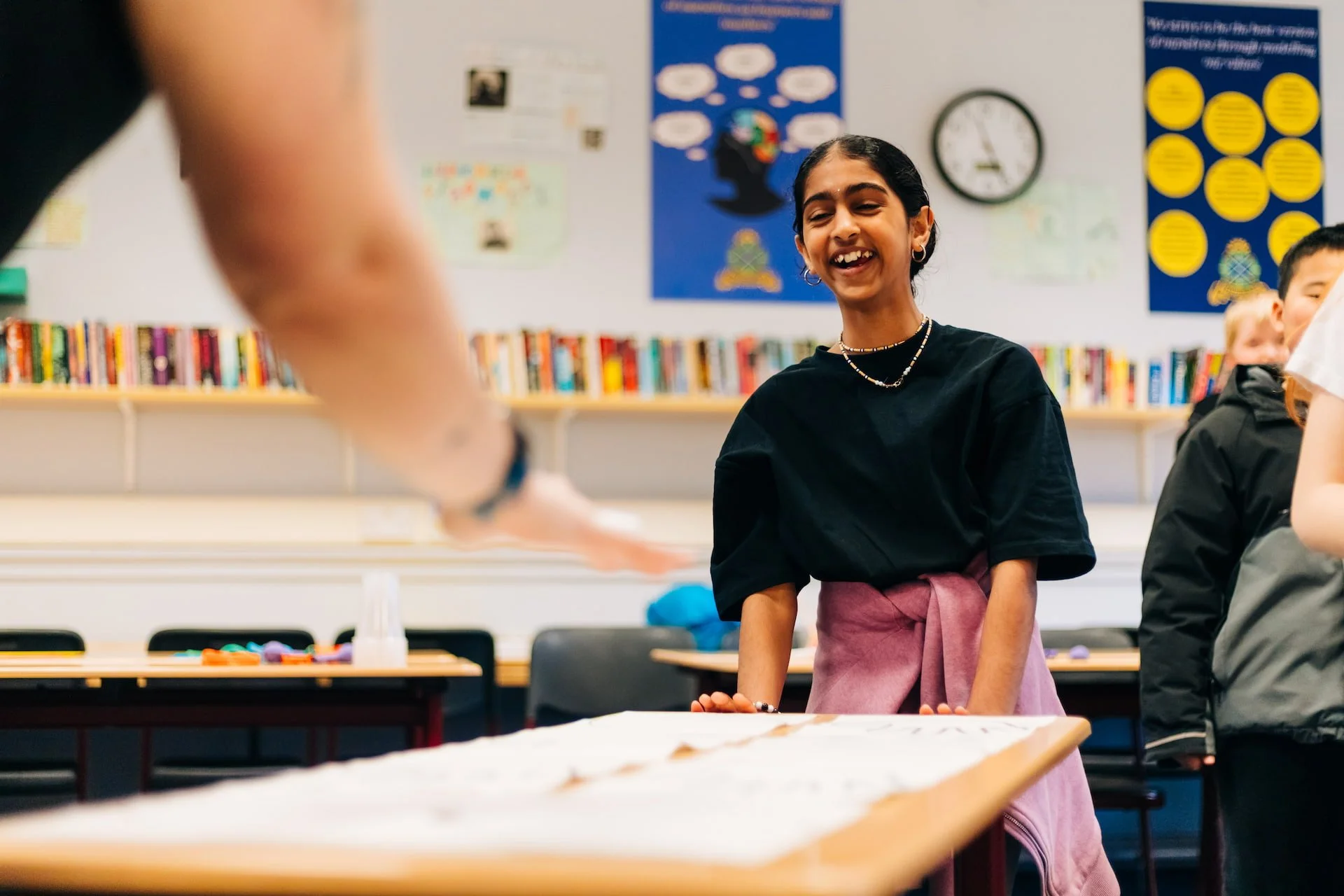 Smiling girl in a black shirt with pink jacket tied around her waist, standing in a classroom with bookshelves and educational posters in the background, looking at an object on the table.