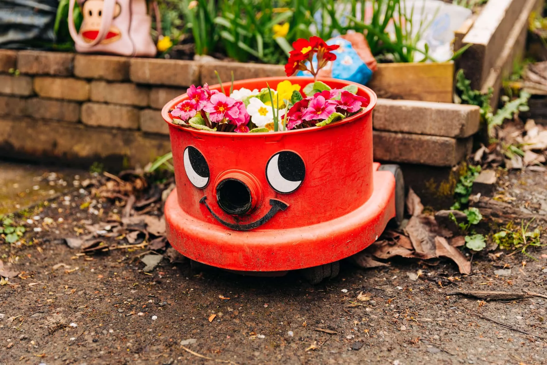 A red flower pot with a smiling face design, filled with colorful flowers, placed on the ground in a garden.