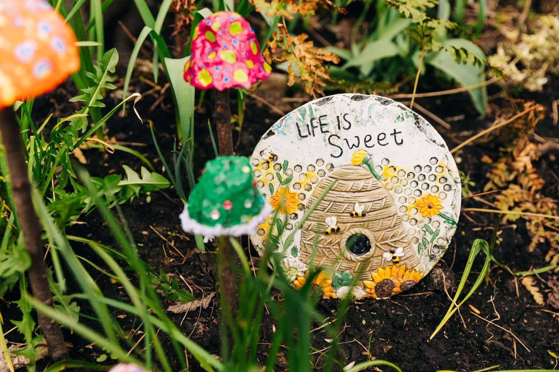 Decorative garden ornament with the words "Life is Sweet," featuring a beehive, bees, flowers, and sunflowers, surrounded by colorful mushrooms and green plants.