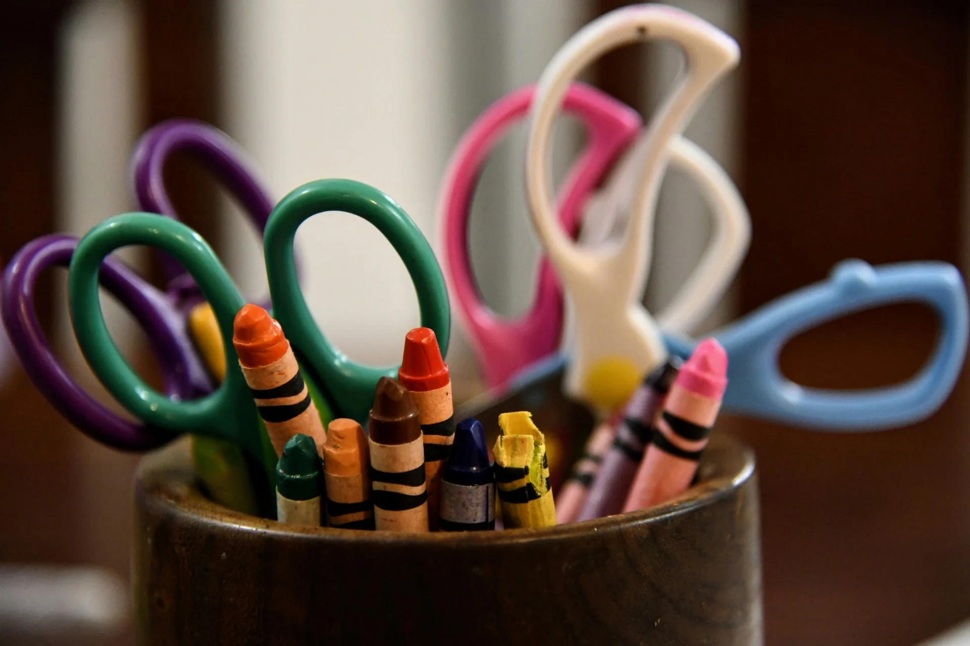 Colorful children's scissors and crayons in a wooden container.