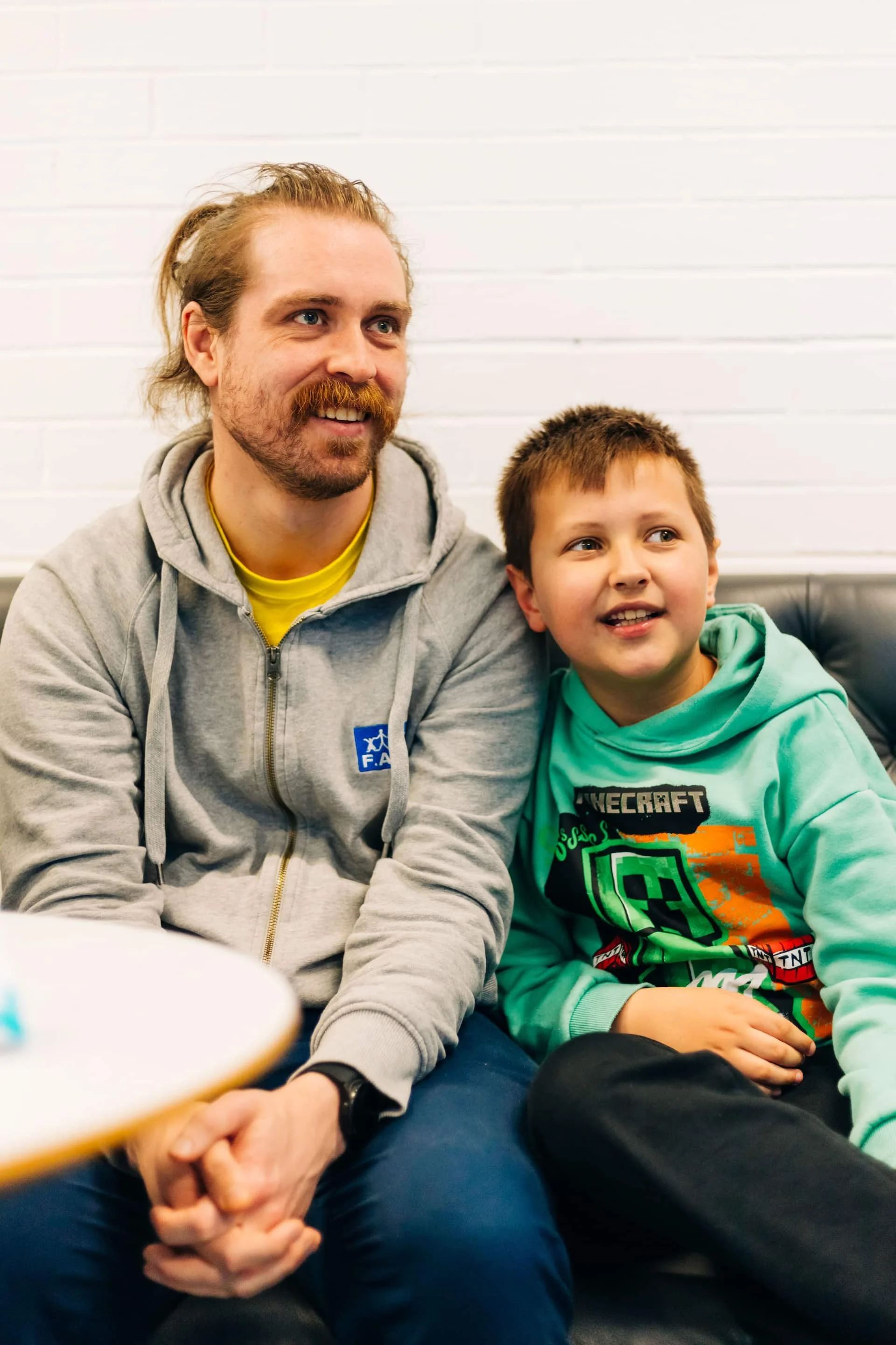 A man with long hair tied back, a beard, and a gray hoodie sitting on a black couch with a boy wearing a Minecraft themed hoodie, both smiling and looking to the side against a white brick wall.