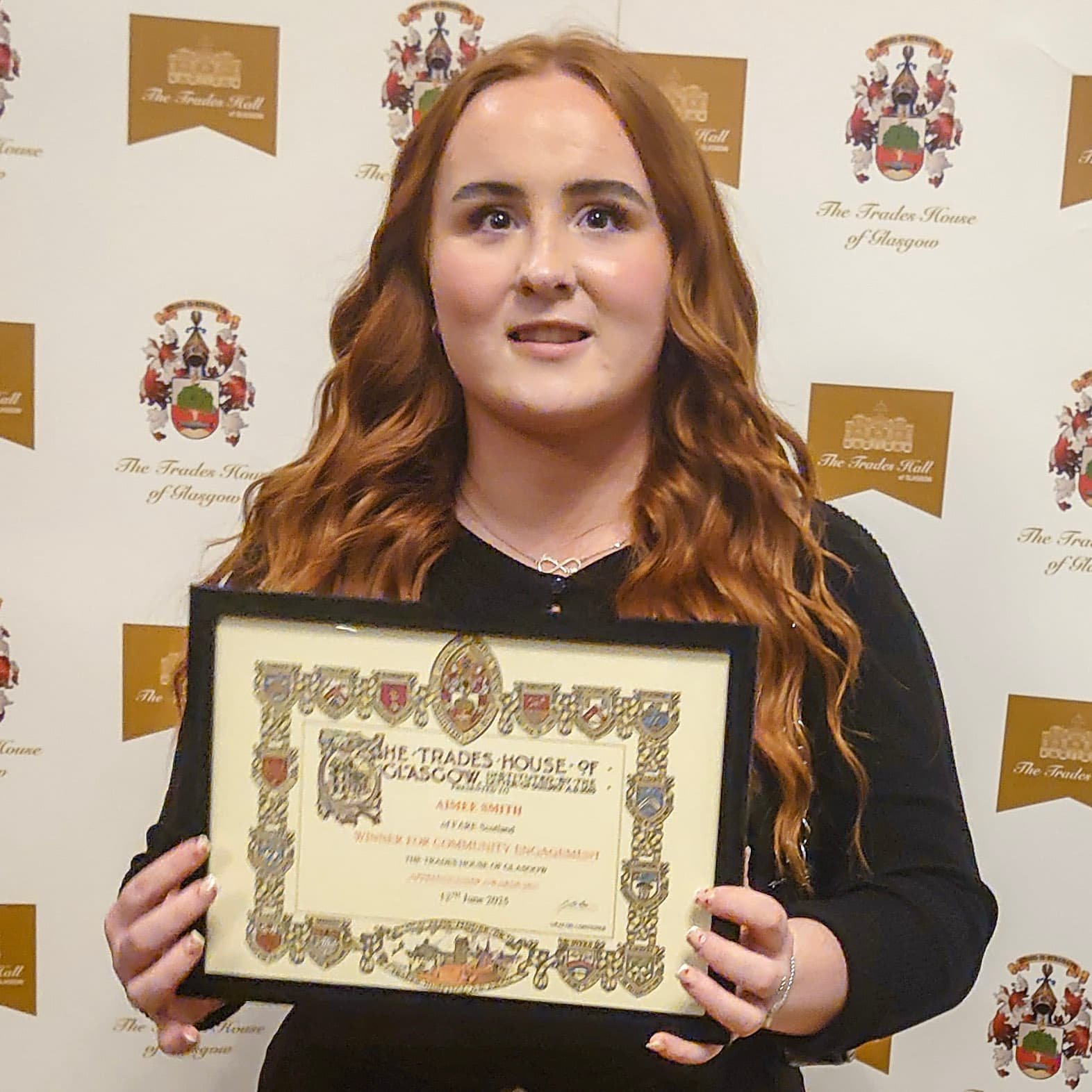 A young woman with long red hair holding a framed certificate at an event at The Trades House of Glasgow, with a backdrop featuring the organization's crest and logos.