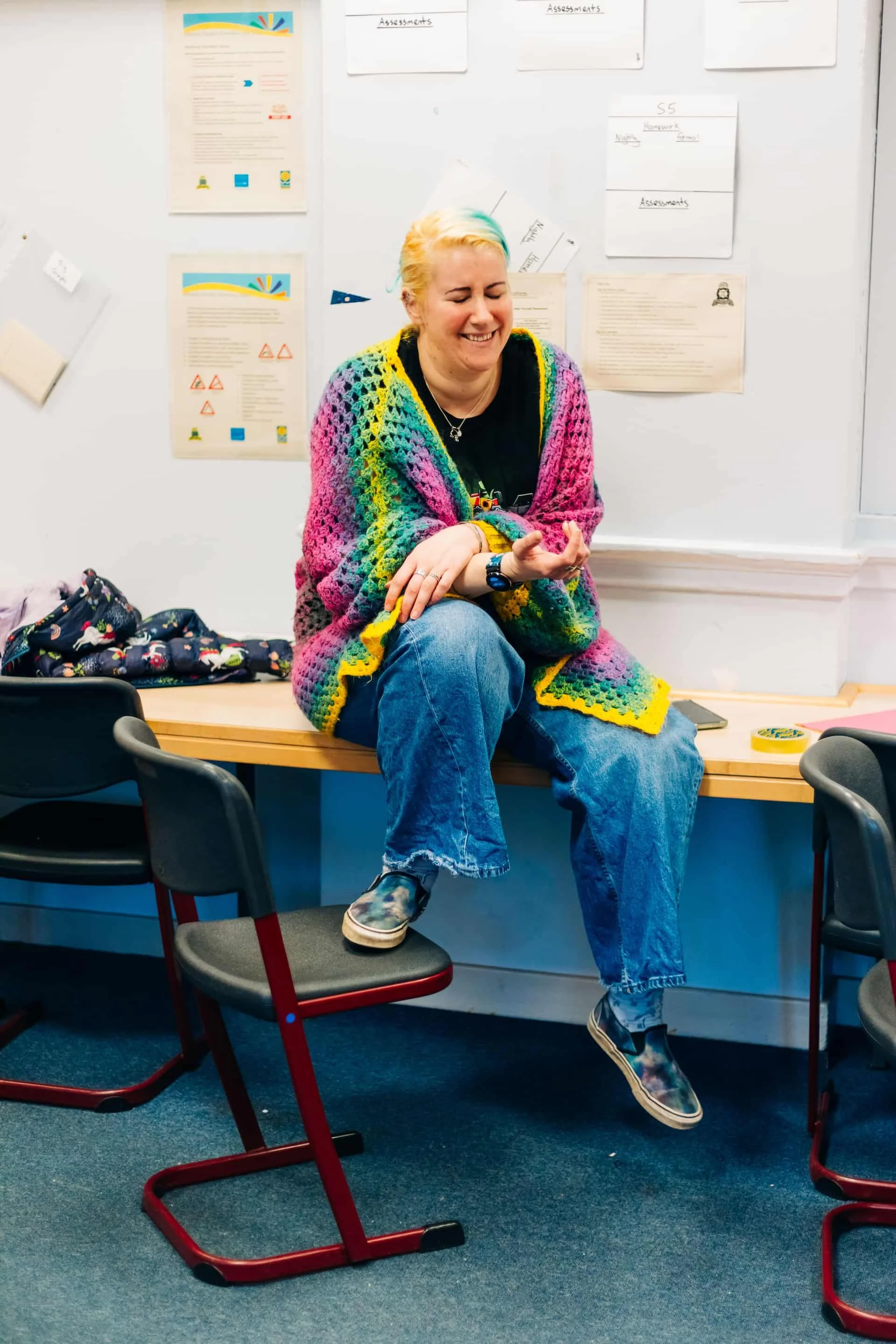 A woman with blonde hair and colorful highlights, wearing a rainbow crochet shawl, sits on a wooden table in a classroom, smiling and enjoying herself. There are chairs, posters, and papers on the wall behind her.