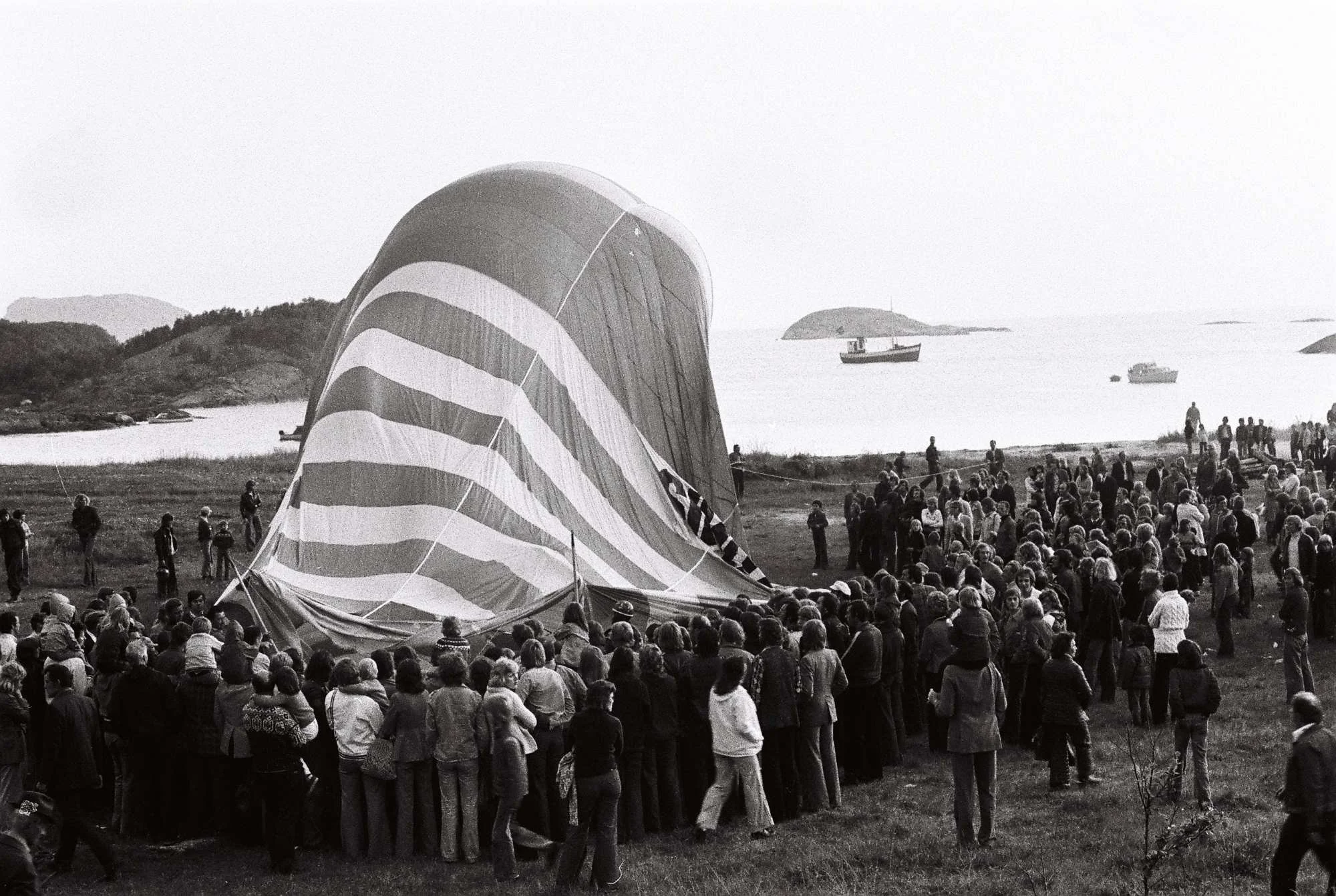 Foto er tatt i Ausvika utenfor Bodø på nordsia av byen i 1975, og viser klargjøring av ballongoppstigning i regi av Bodø Ballong Club. På bildet sert vi masse mennesker som har møtt opp for å overvære begivenheten.