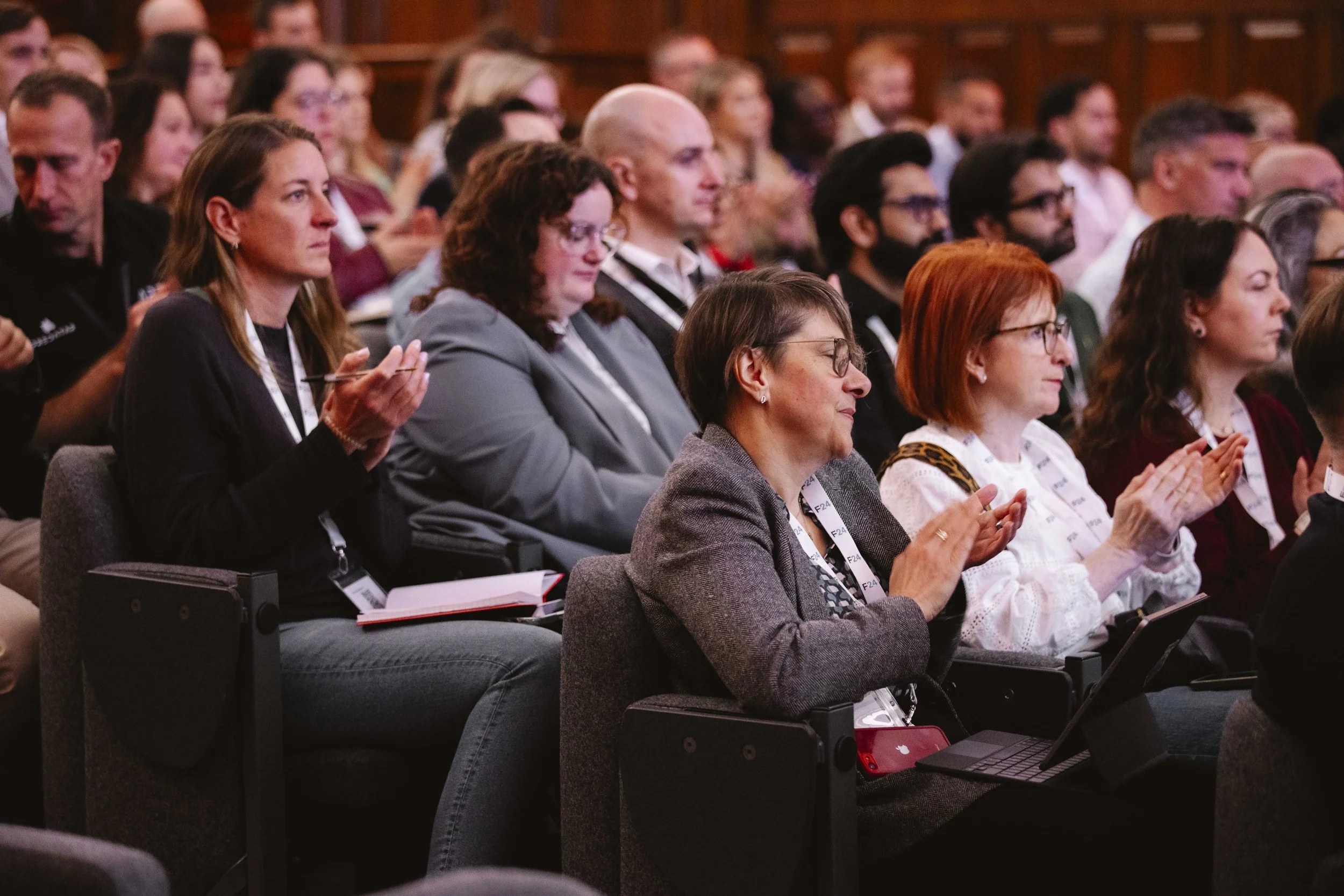 Audience members sitting and clapping at a conference or seminar, with some taking notes or using electronic devices.