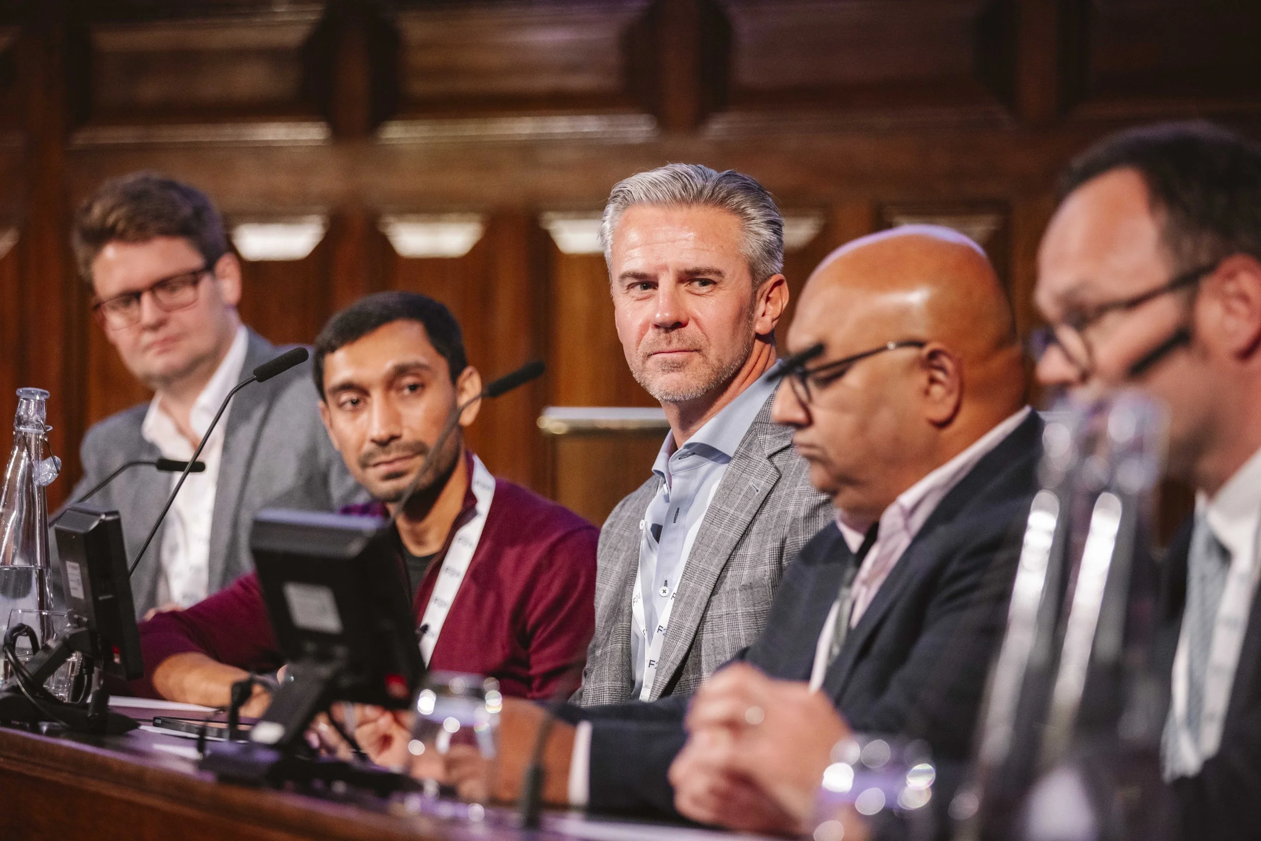 A group of five men sitting at a conference or panel discussion, with microphones in front of them, in a wood-paneled room.