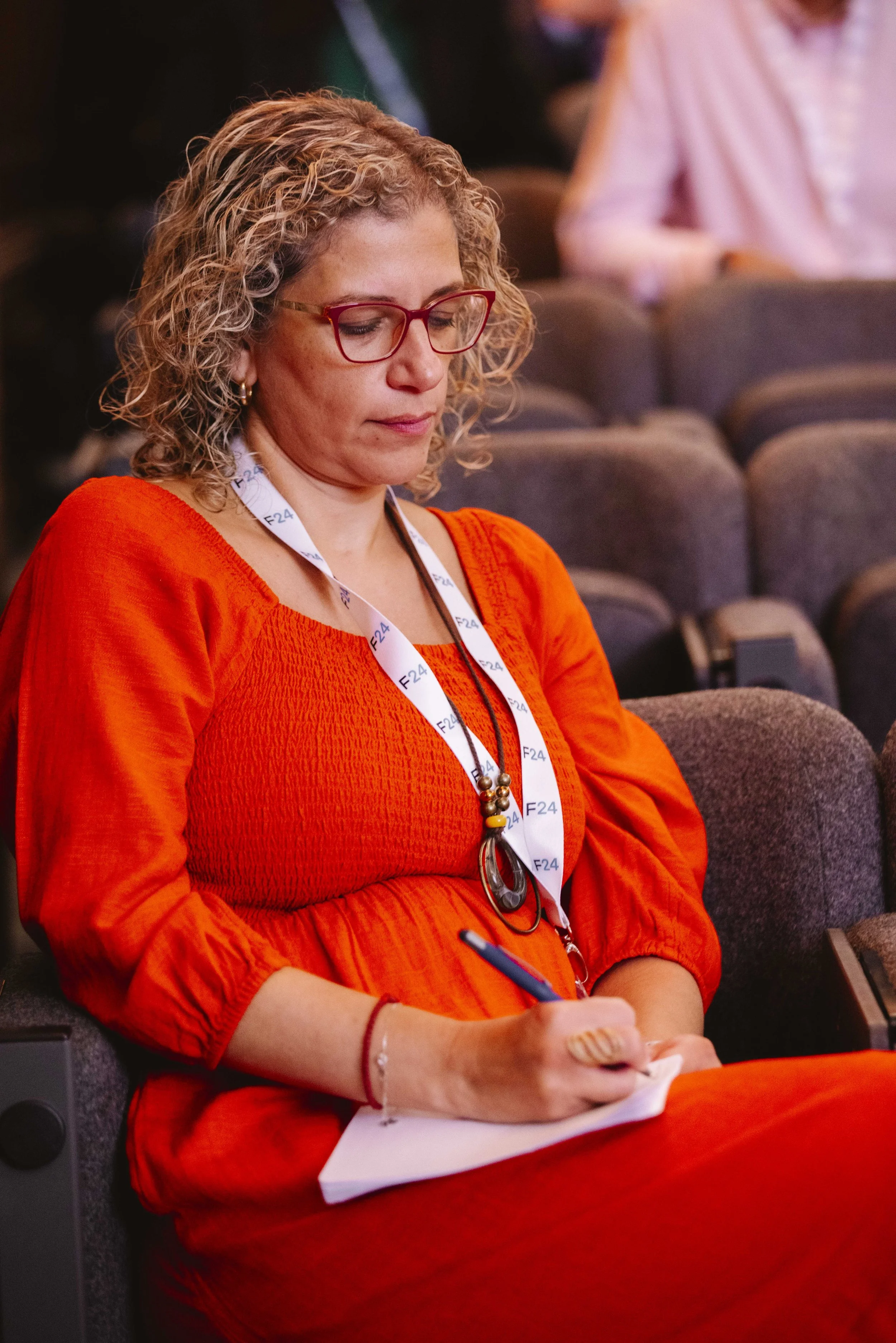 A woman with curly blonde hair, wearing red glasses and an orange dress, sitting in a conference hall taking notes with a pen and notepad.