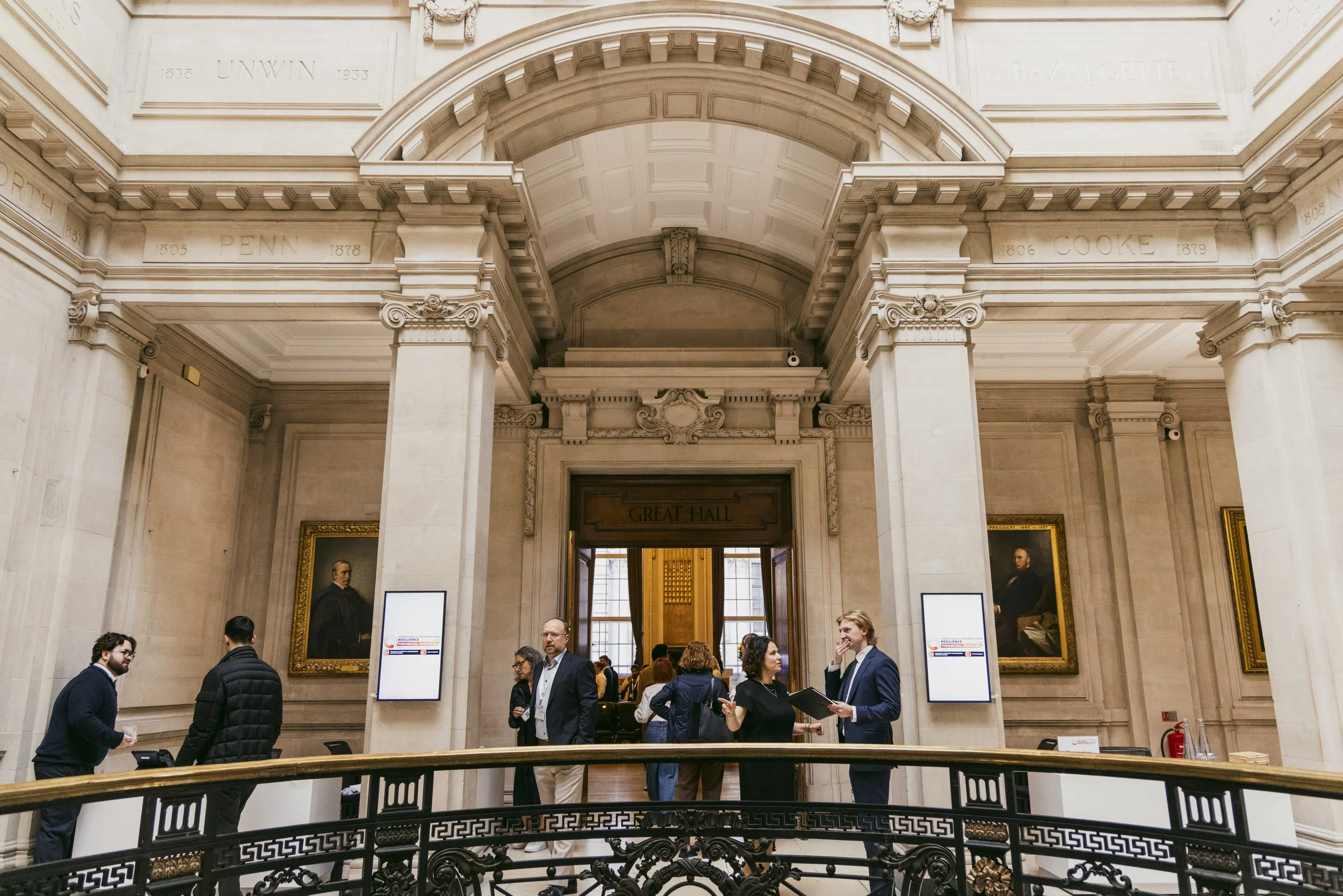 People standing and conversing inside a historic building with grand architectural details, portrait paintings, and digital screens, near the entrance to a room labeled "GREAT HALL."