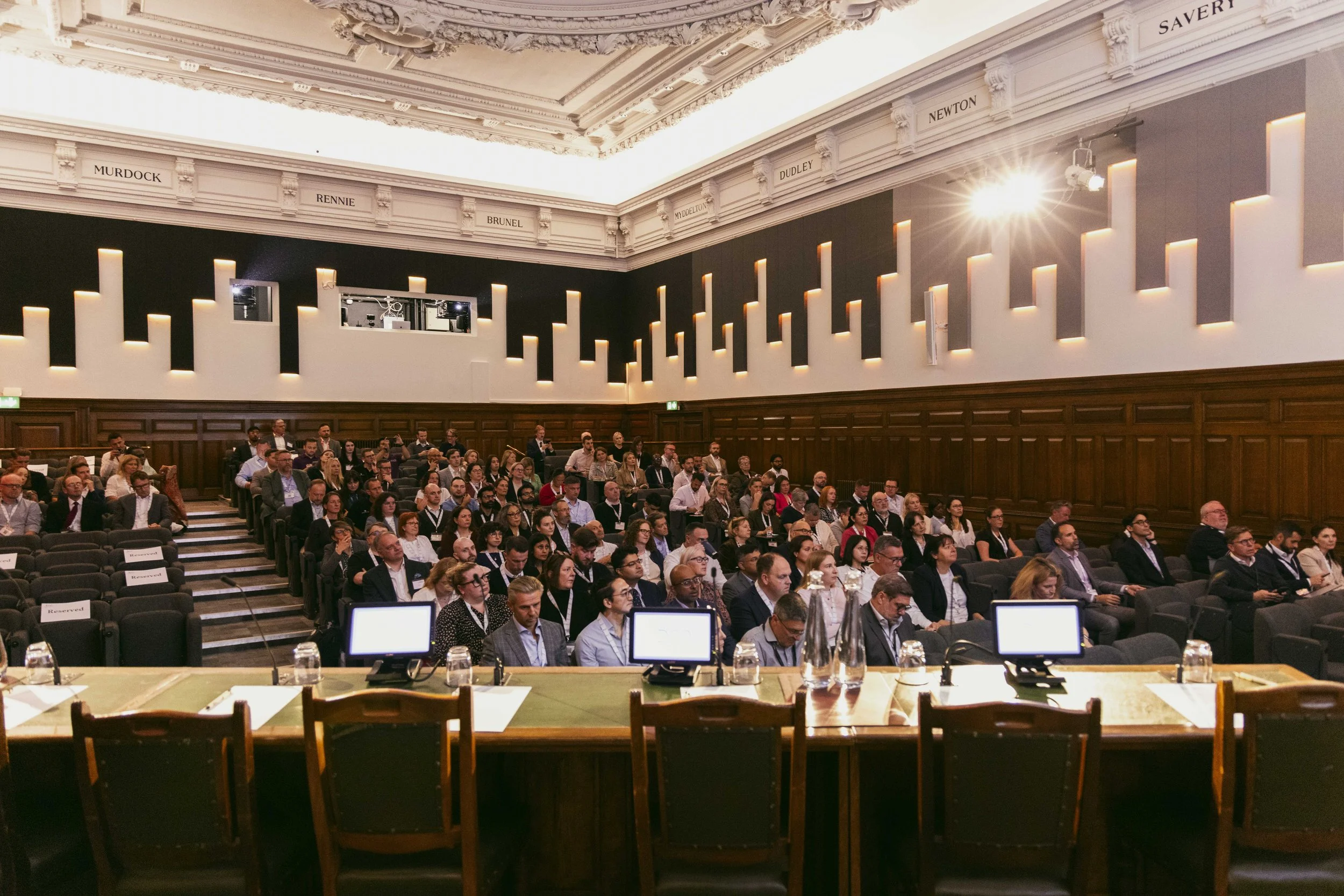Business conference in a large, historic room with high ceiling and ornate decorations. Audience seated in rows, facing the front, with tables and digital screens at the speaker's area.
