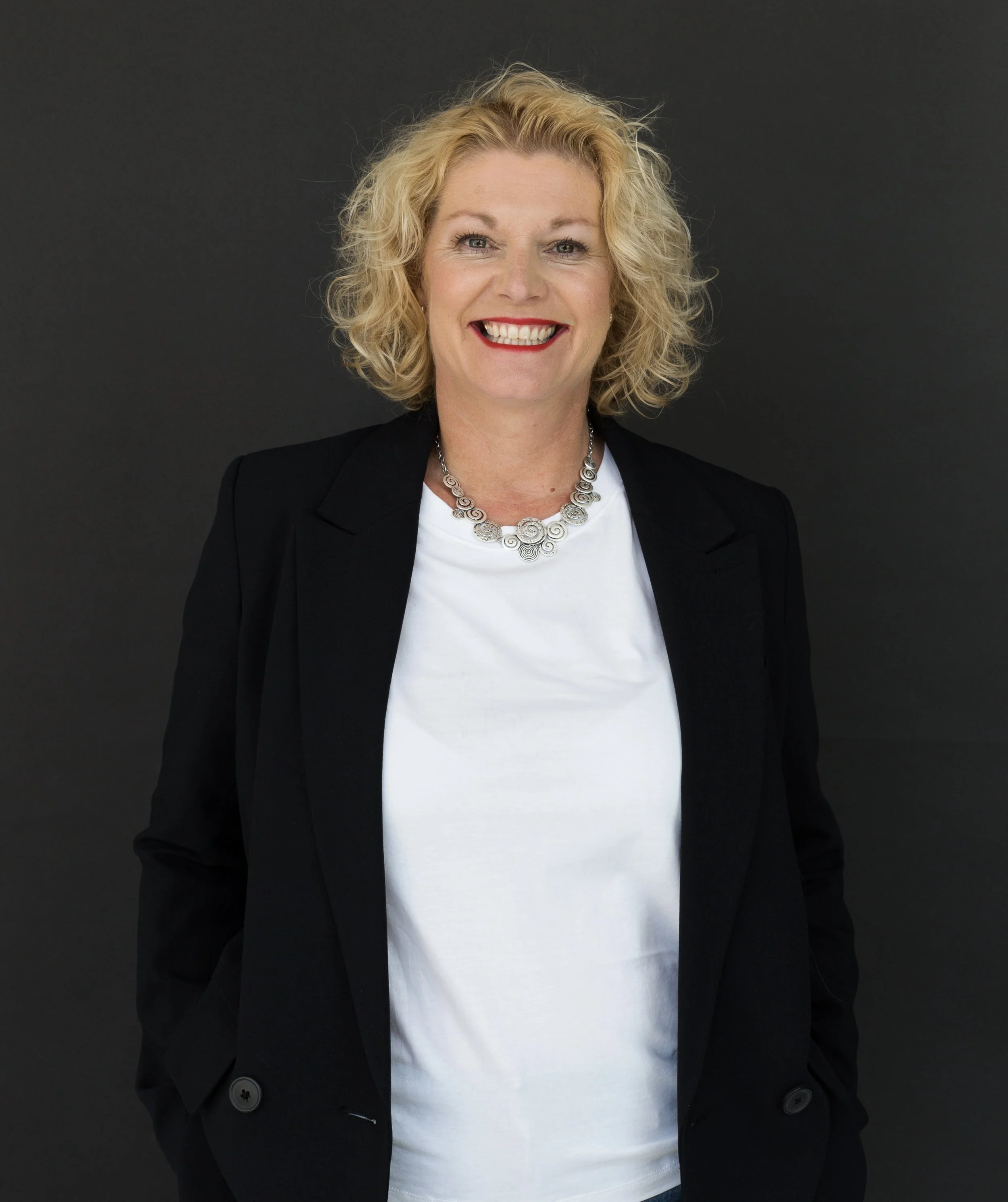 Smiling woman with curly blonde hair wearing a black blazer, white shirt, and a silver necklace, standing against a dark background.