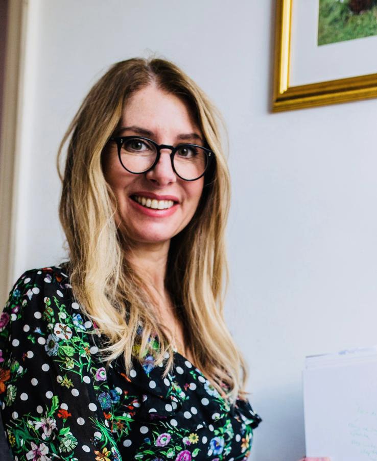 Smiling woman with glasses and long blonde hair, wearing a black floral dress, standing indoors next to a picture frame.