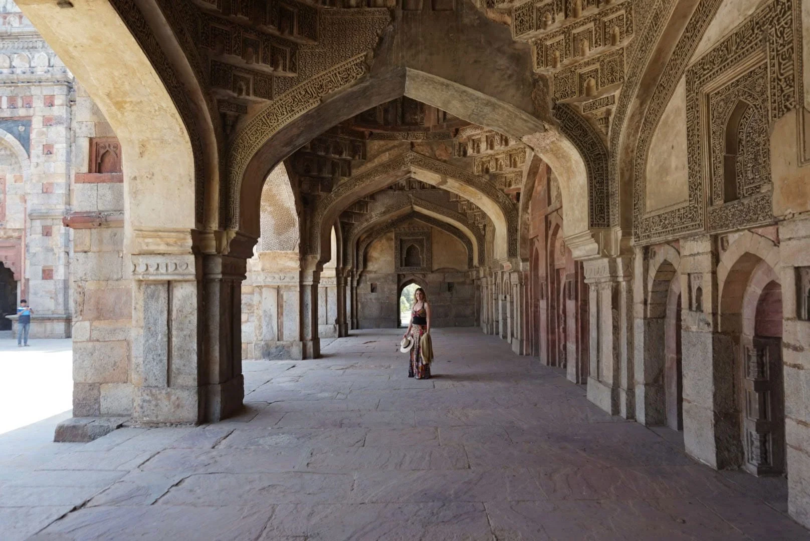 A woman dressed in a long, colorful skirt and a black top, holding a hat and a bag, stands in an ancient arched corridor with intricate carvings on the walls and ceiling. The corridor appears to be part of a historic monument or palace, with sunlight illuminating the scene from the far end.