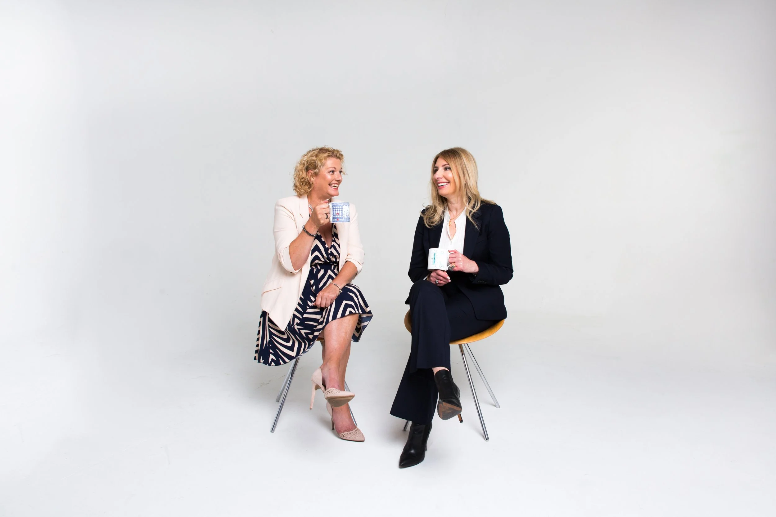 Two women sitting on chairs, smiling and holding mugs, engaged in conversation against a plain white background.