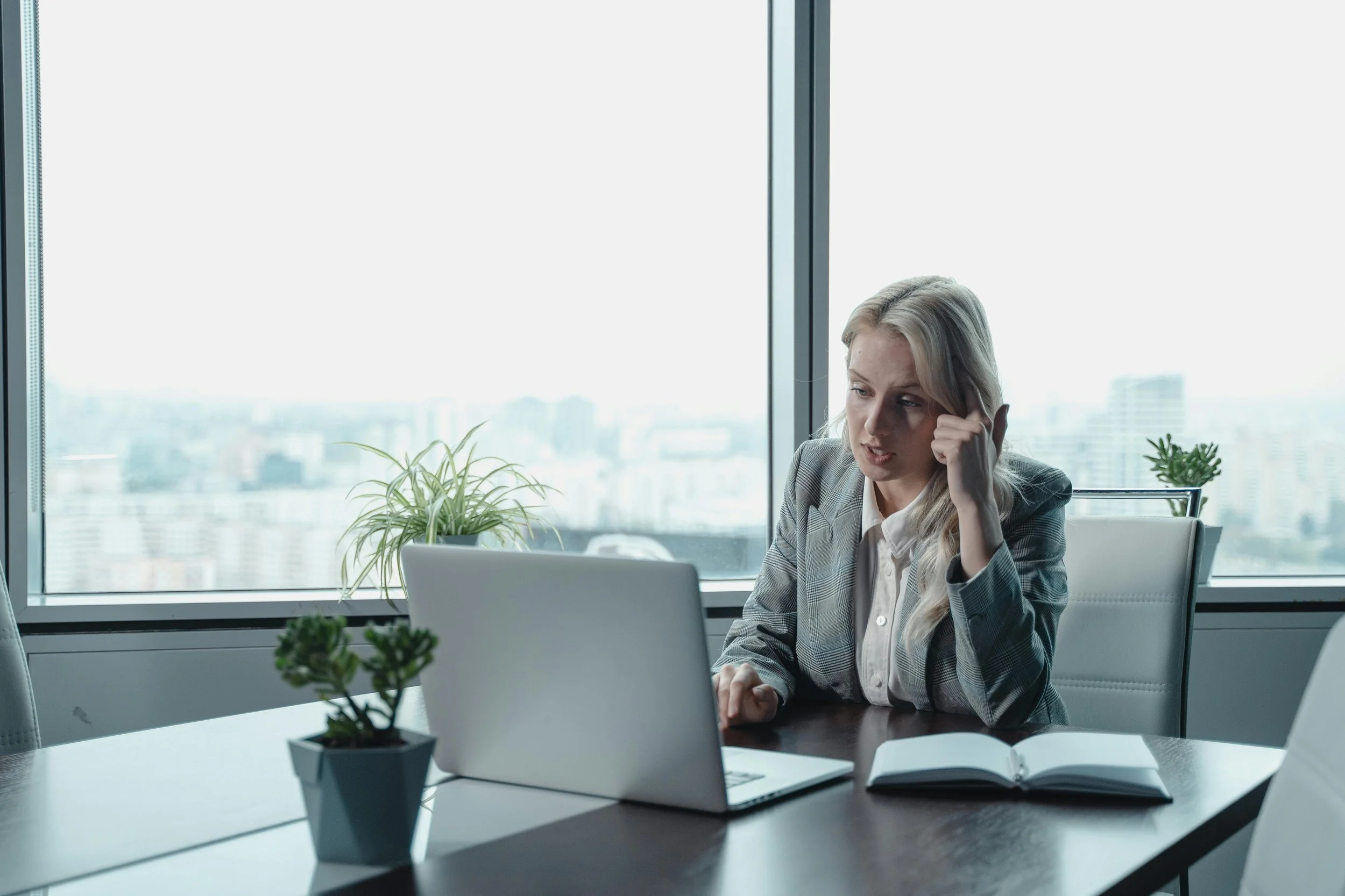 A woman with blonde hair working on a laptop at a desk in a modern office near a large window with a city view. There are two potted plants on the desk and an open book.