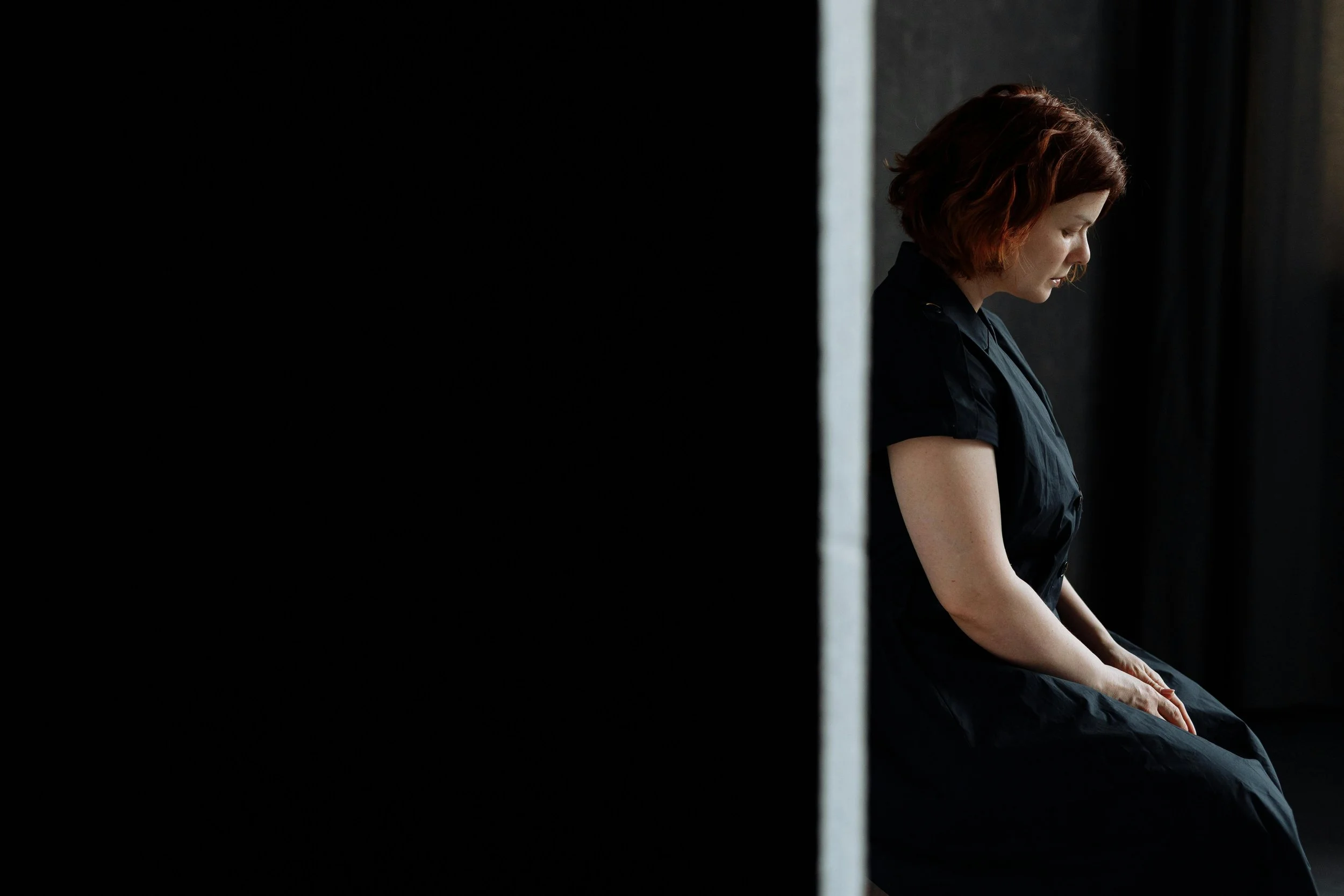 A young woman with short red hair sits alone in a dark room, her head bowed and eyes closed, wearing a black dress.