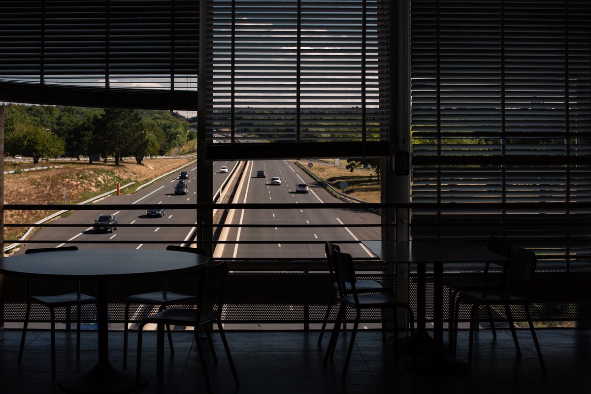 Vue de l'extérieur d'une autoroute avec plusieurs voitures, vue partielle d'une salle sombre avec des tables et des chaises devant une grande fenêtre avec des volets roulants.