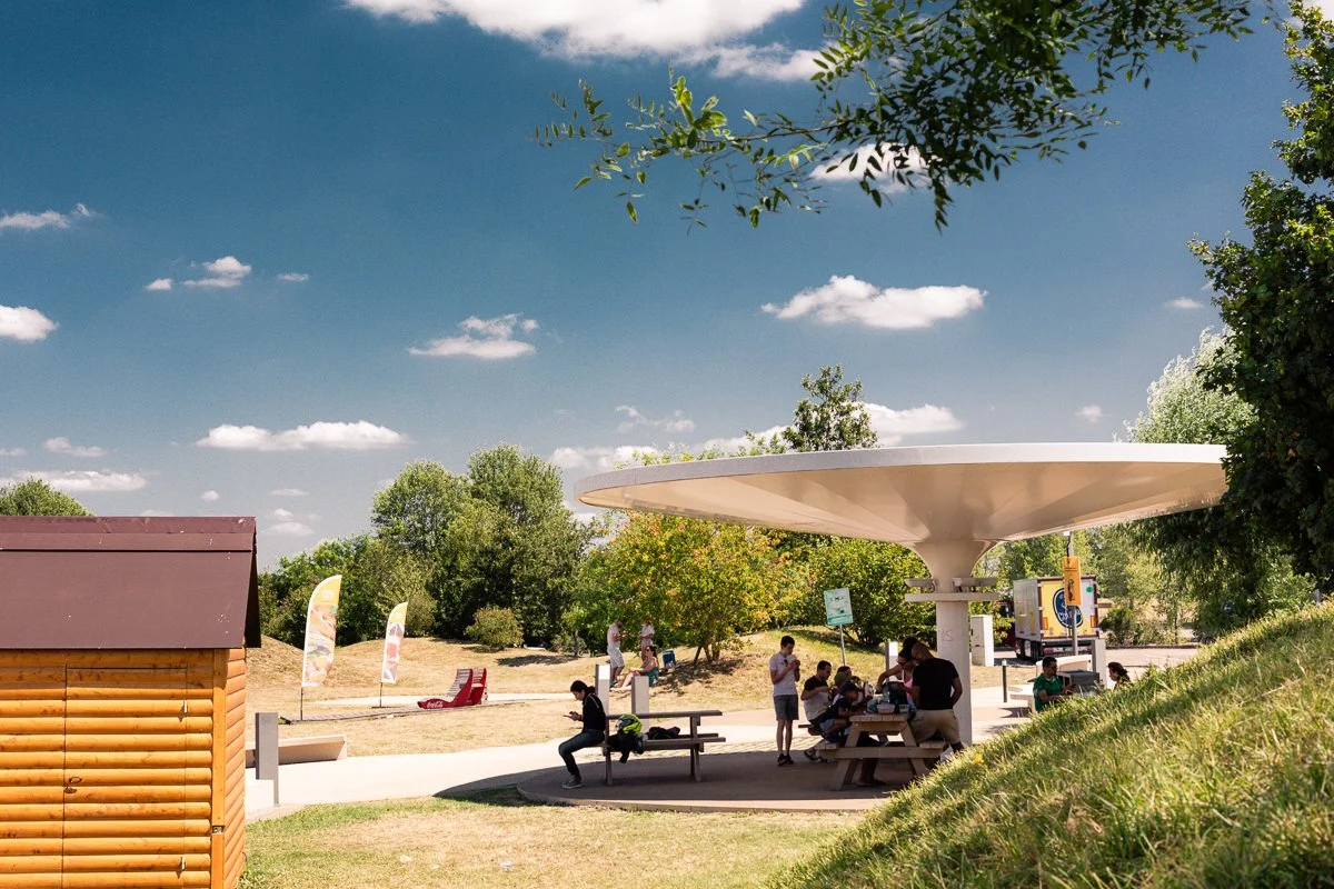 Parc avec une structure abritant des tables, une maison en bois, et des arbres verts sous un ciel bleu avec quelques nuages