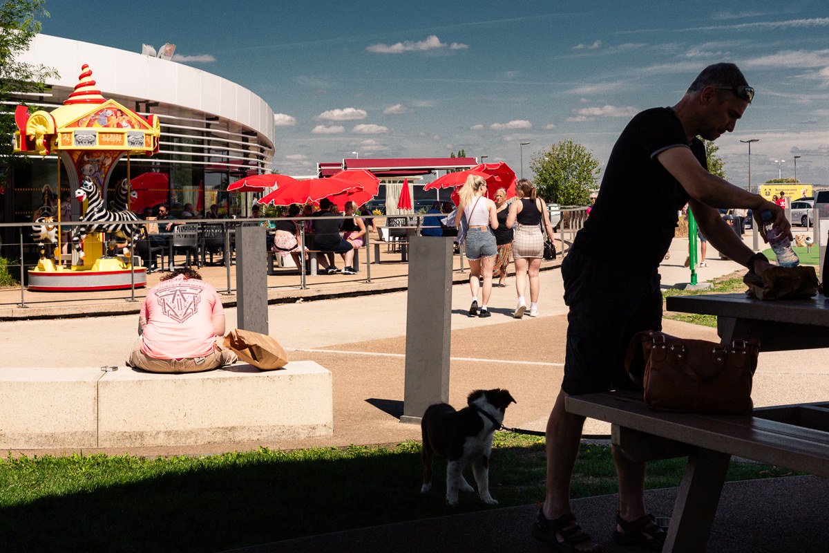 Une personne assise sur un banc, un homme préparant quelque chose sur une table de pique-nique, un chien noir et blanc à ses côtés, un groupe de personnes discutant sous des parasols rouges, un manège à chevaux de bois, un bâtiment moderne en arrière-plan, le tout par une journée ensoleillée.