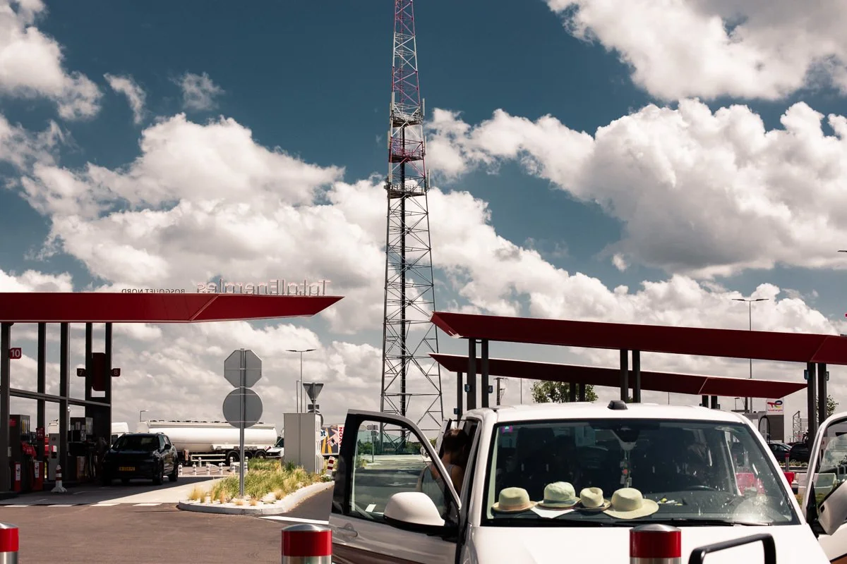 Une station d'essence avec des voitures garées, des chapeaux posés sur le tableau de bord d'une voiture blanche, un grand pylône métallique et un ciel avec des nuages blancs et un ciel bleu.