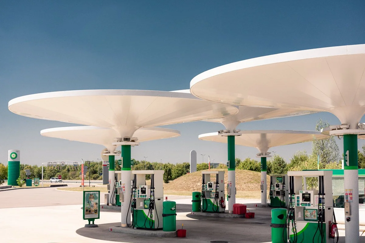 Station-service BP avec plusieurs pompes à essence sous des abris modernes en forme de grands disques blancs, entourée d'un environnement naturel ensoleillé avec des arbres et un ciel clair.