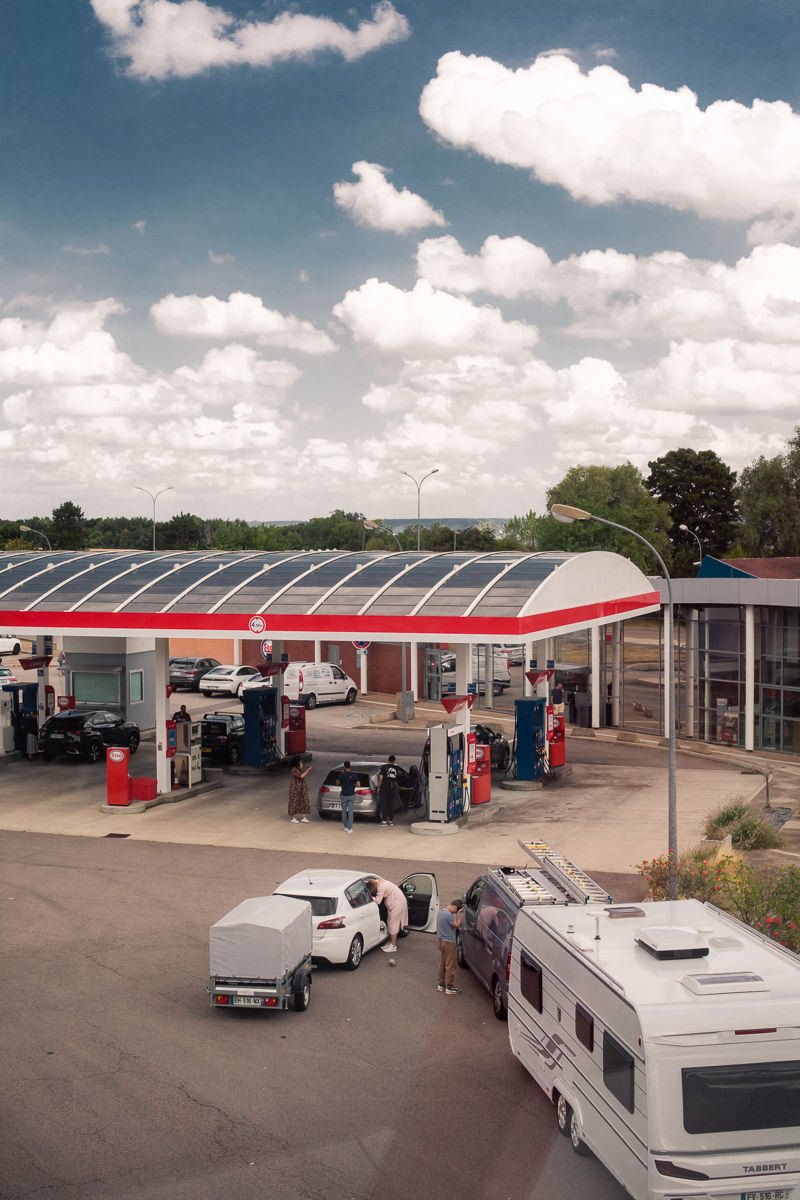 Station-service avec plusieurs voitures en train de faire le plein, quelques personnes autour des véhicules, un camping-car stationné à côté.