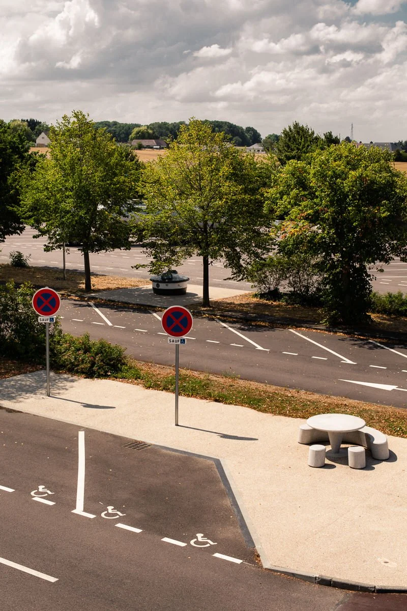 Un parking avec des places réservées aux personnes handicapées, une table de pique-nique en béton dans un espace vert, et des panneaux interdisant le stationnement sauf pour certains usagers. En arrière-plan, des arbres et une route.
