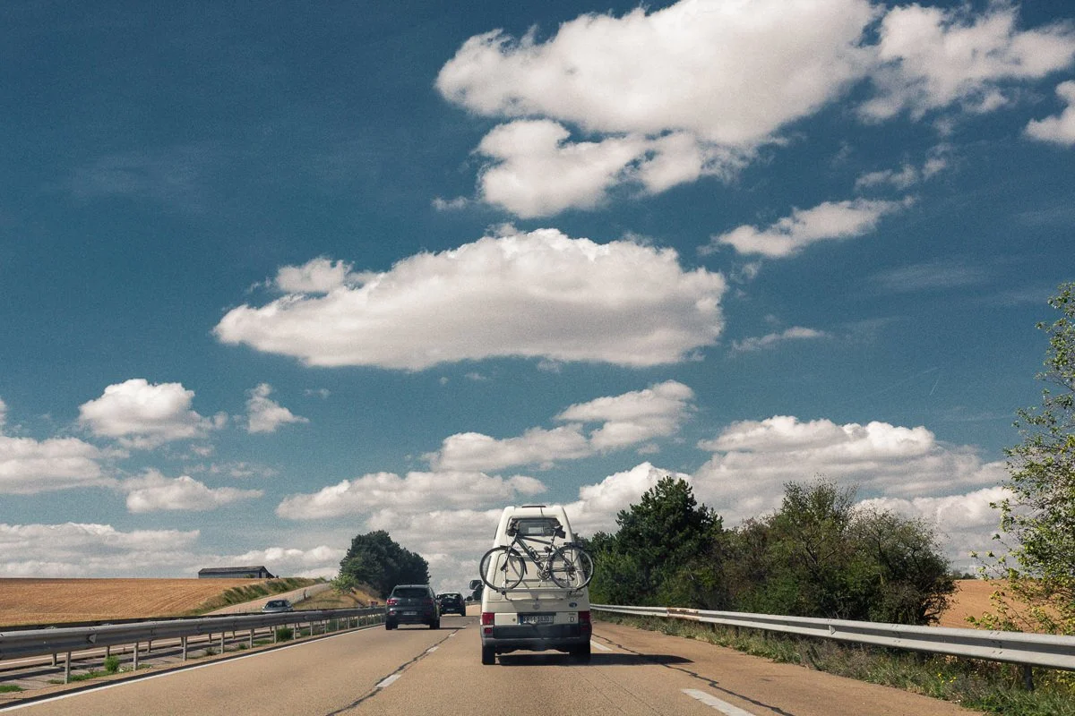 Une voiture avec un vélo attaché à l'arrière conduit sur une route en plein air par une journée ensoleillée, avec un ciel bleu et des nuages blancs.