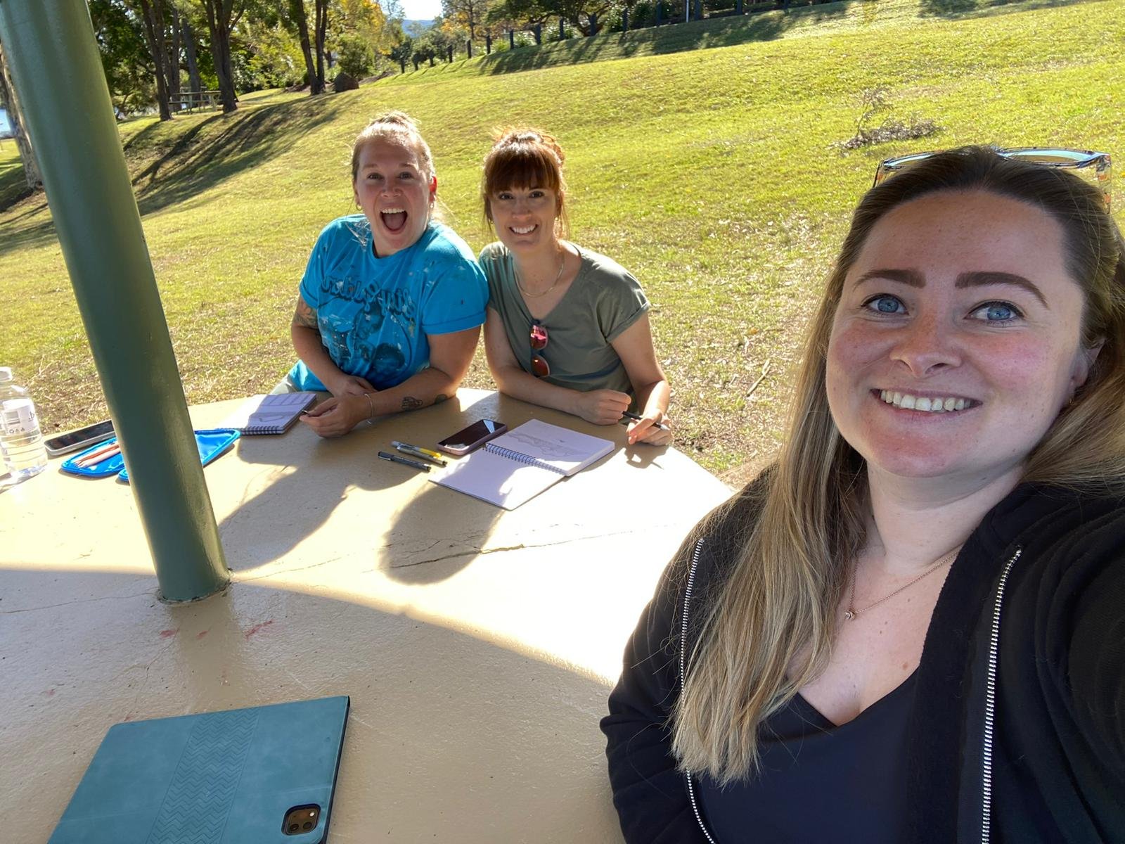 three women sat around a round bench outside in the sunshine posing for a photo