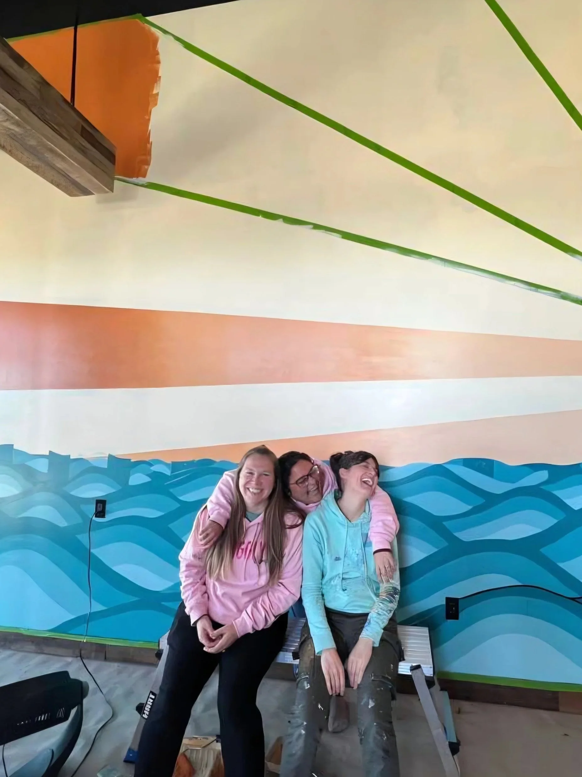 Jess, Chris and Kate all mural artists laughing out loud whilst sat on a bench in front of their mural, trying to take a photo.