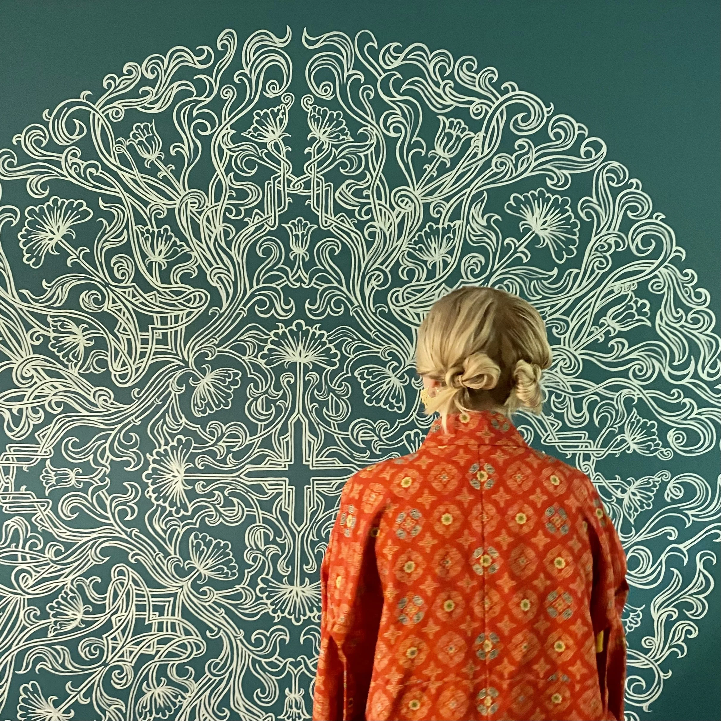 Elizabeth stands facing her wall mural. with a read patterned coat and two buns in her hair