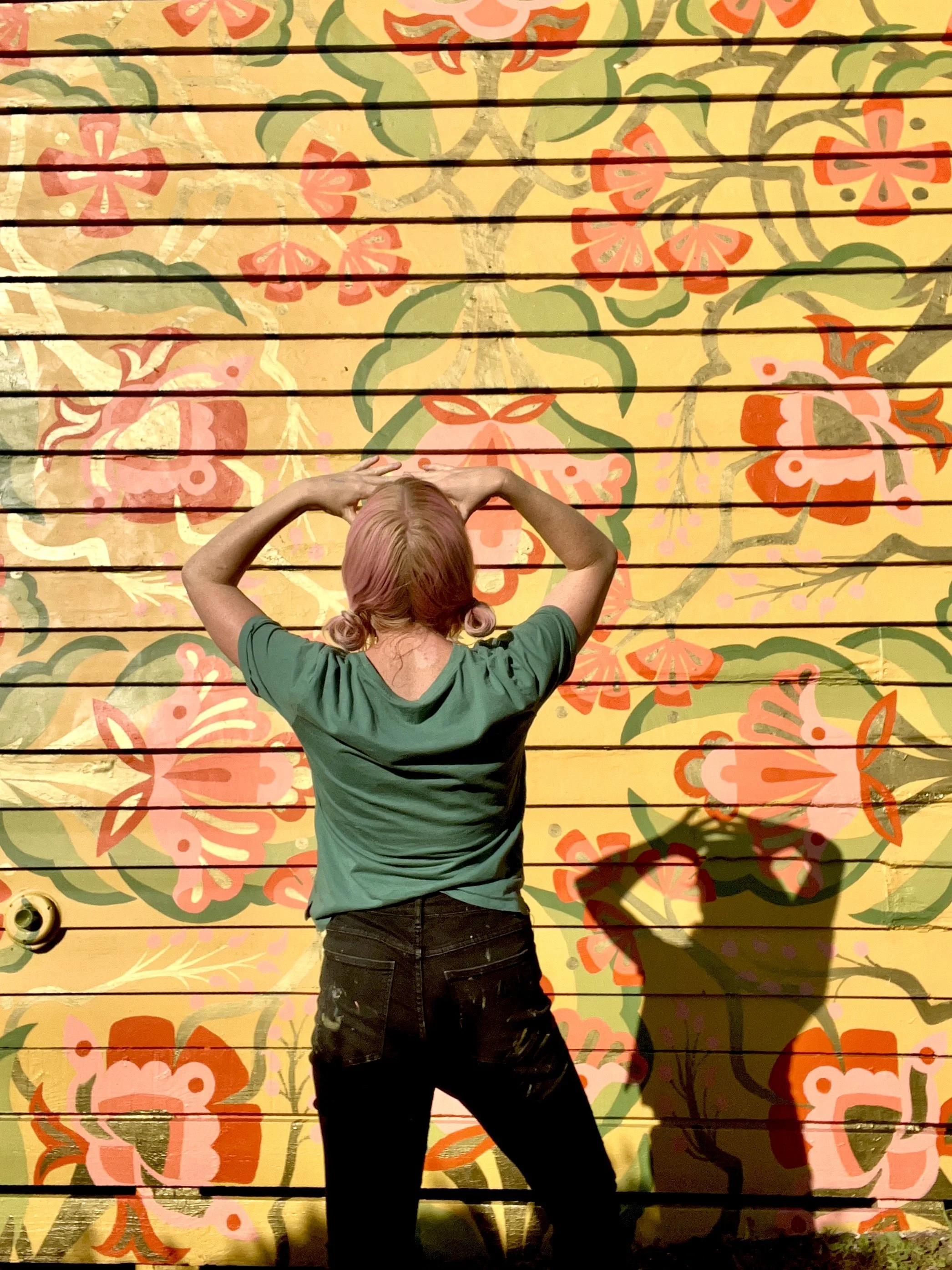 Elizabeth standing in front of her mural. Bright yellow floral design sunlight streaming onto the wooden wall.