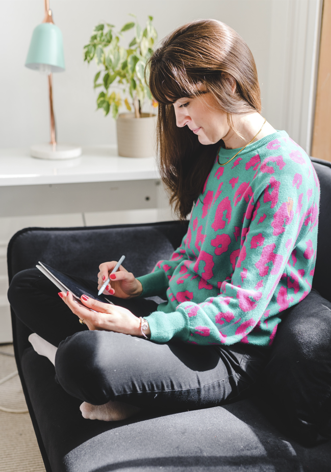 a woman sat cross legged on the sofa with her laptop. Wearing a turquois and fuchsia pink leopard print jumper