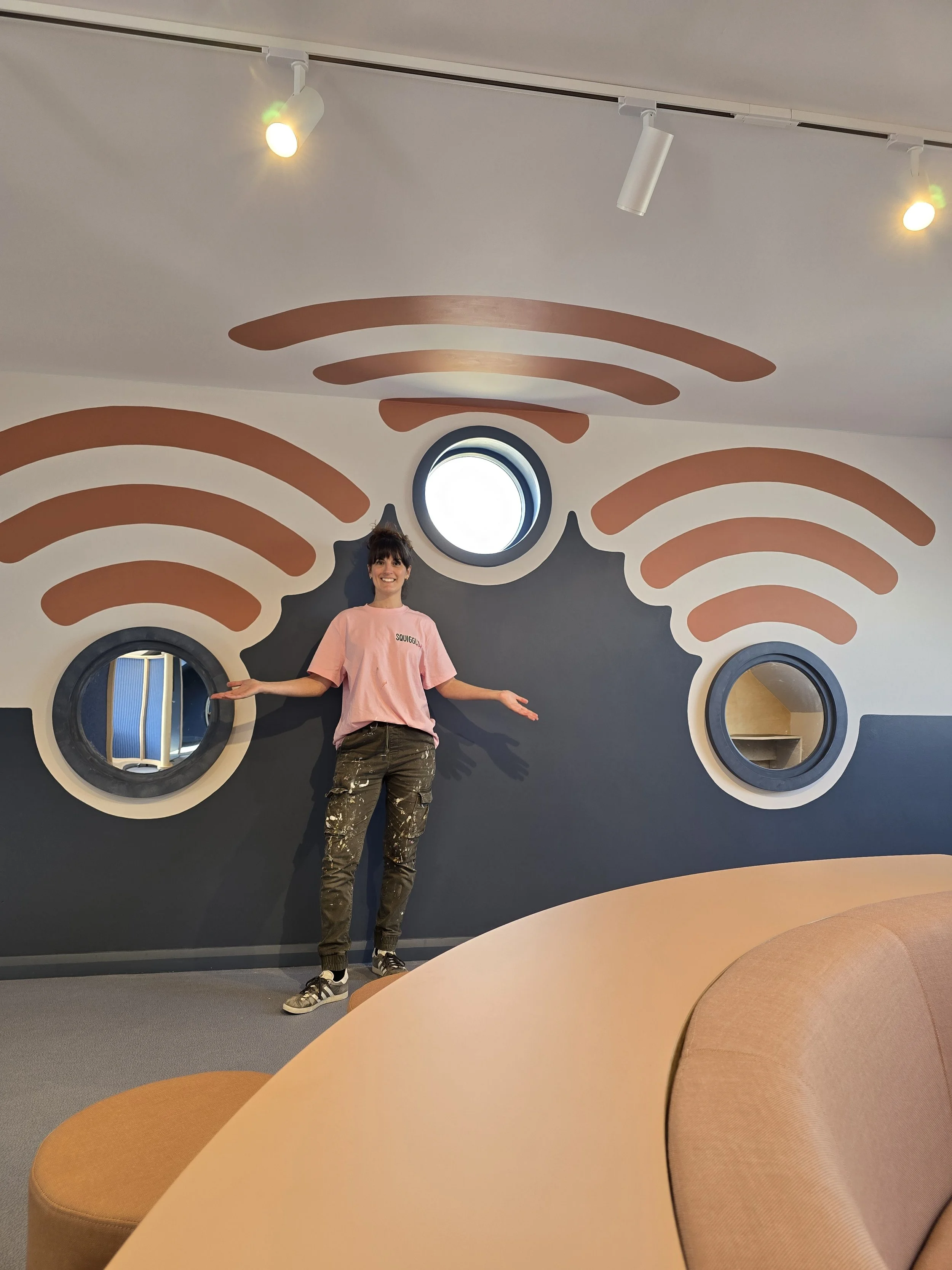 kate the artist stands in front of a finished mural. orange wifi signals radiate out from a blue wall with round porthole windows, for a new IT suite at a local school.