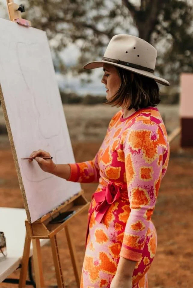 The pink galah mural artist stood sketching at an easel in the outback of Australia. Wearing a cowboy hat, boots and a beautiful floral dress.