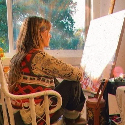 Photo of a young female artist sat on a wicker chair near the window. Light streaming through whilst she stares at her artwork on canvas surrounded by paint and brushes