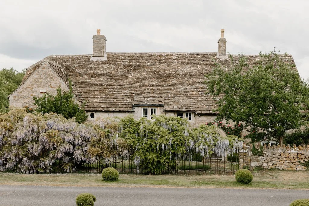 A stone cottage with a tiled roof, white window frames, and surrounded by flowering bushes and trees.