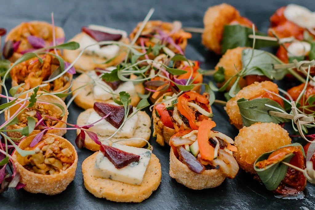 Assorted appetizer bites including mini croquettes, topped crostinis with cheese and cured meat, and vegetable-topped bites garnished with microgreens on a black platter.