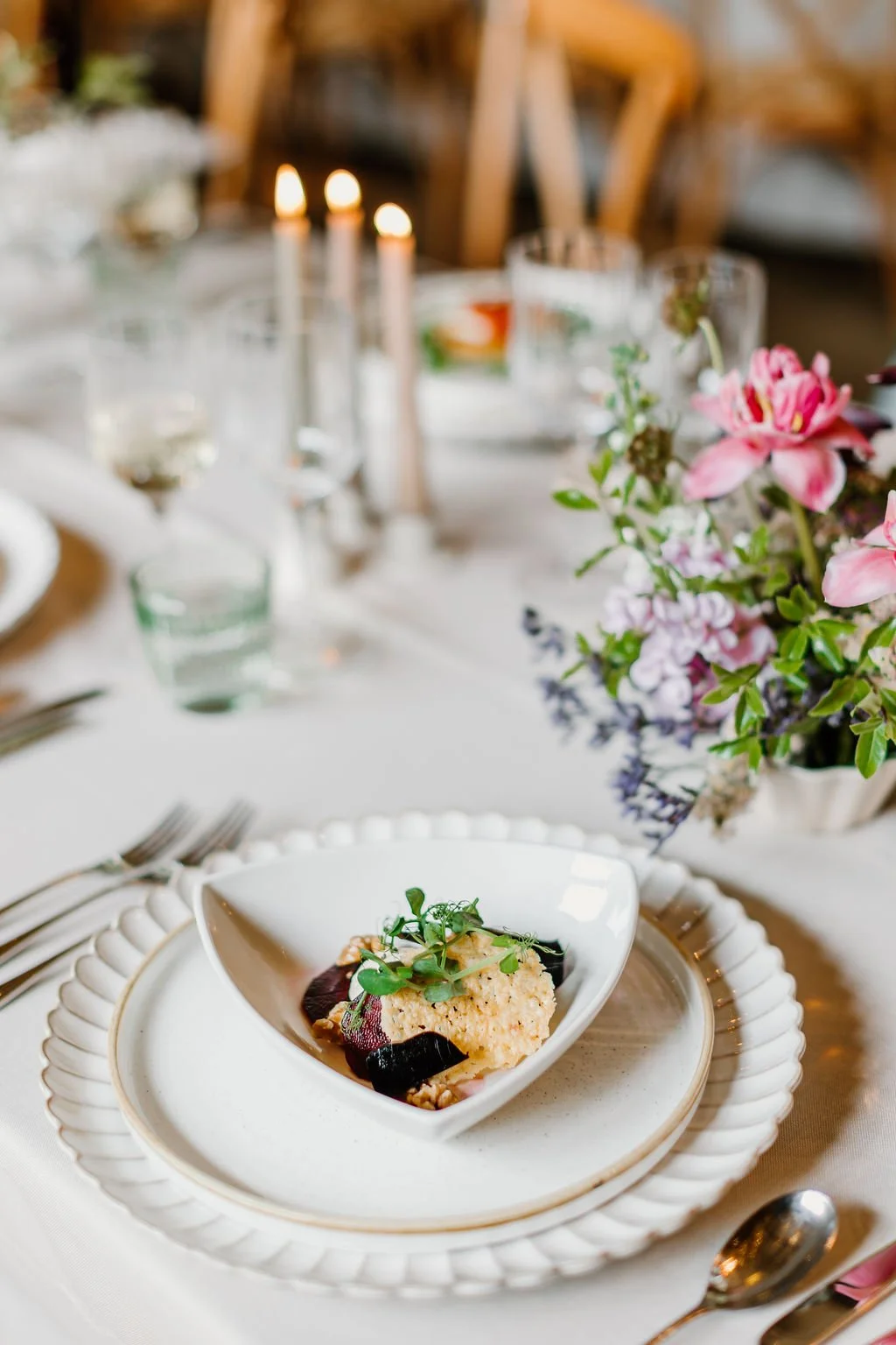 Elegant table setting with a white dish containing a gourmet appetizer topped with microgreens, on a white plate with a scalloped edge, surrounded by floral centerpiece and candles.
