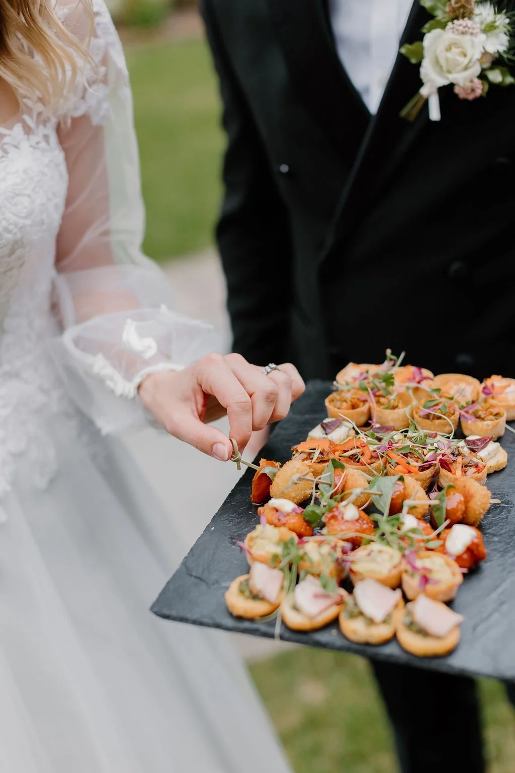 Close-up of a bride and groom at a wedding, with the groom serving appetizer bites from a black platter. The bride is wearing a white lace wedding dress with sheer, embellished sleeves. The platter holds small, topped savory snacks garnished with mic