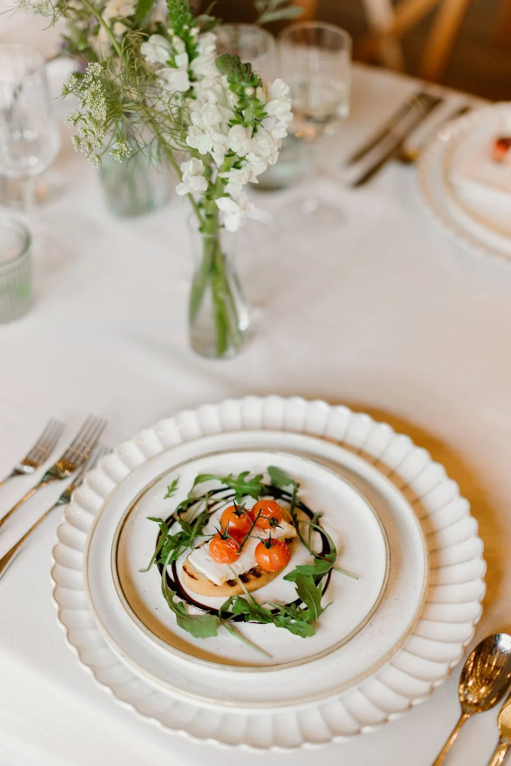 Elegant table setting with a white ceramic plate featuring a grilled dish topped with cherry tomatoes, arugula, and cheese, surrounded by a white scalloped charger, gold flatware, and a flower arrangement with white flowers and greenery in a glass va
