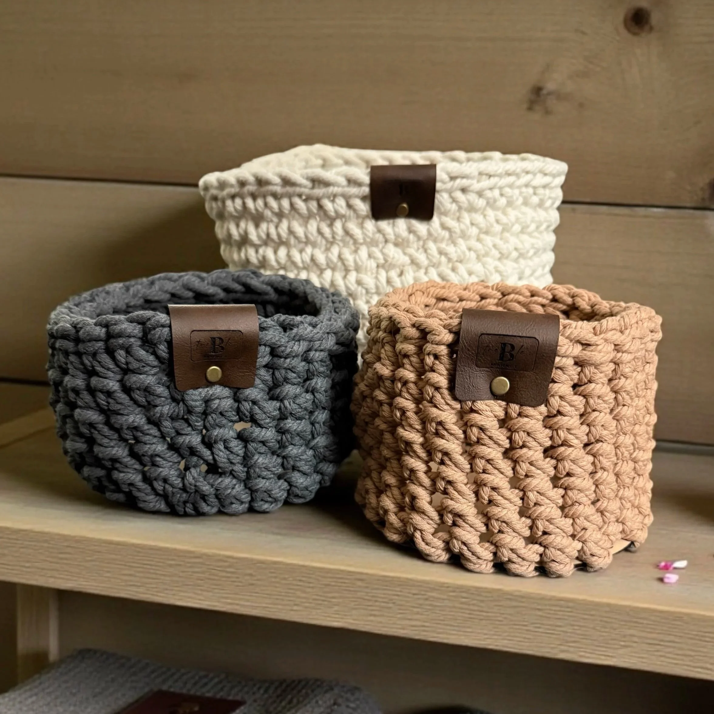 Three chunky knit storage baskets in cream, gray, and peach colors on a wooden shelf.